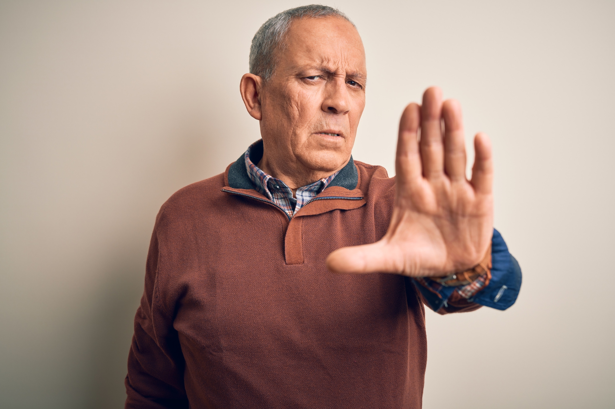 An older man with short gray hair is standing against a plain background. He is wearing a brown sweater over a checkered shirt. He has an outstretched hand towards the camera, palm open, and a serious expression on his face.