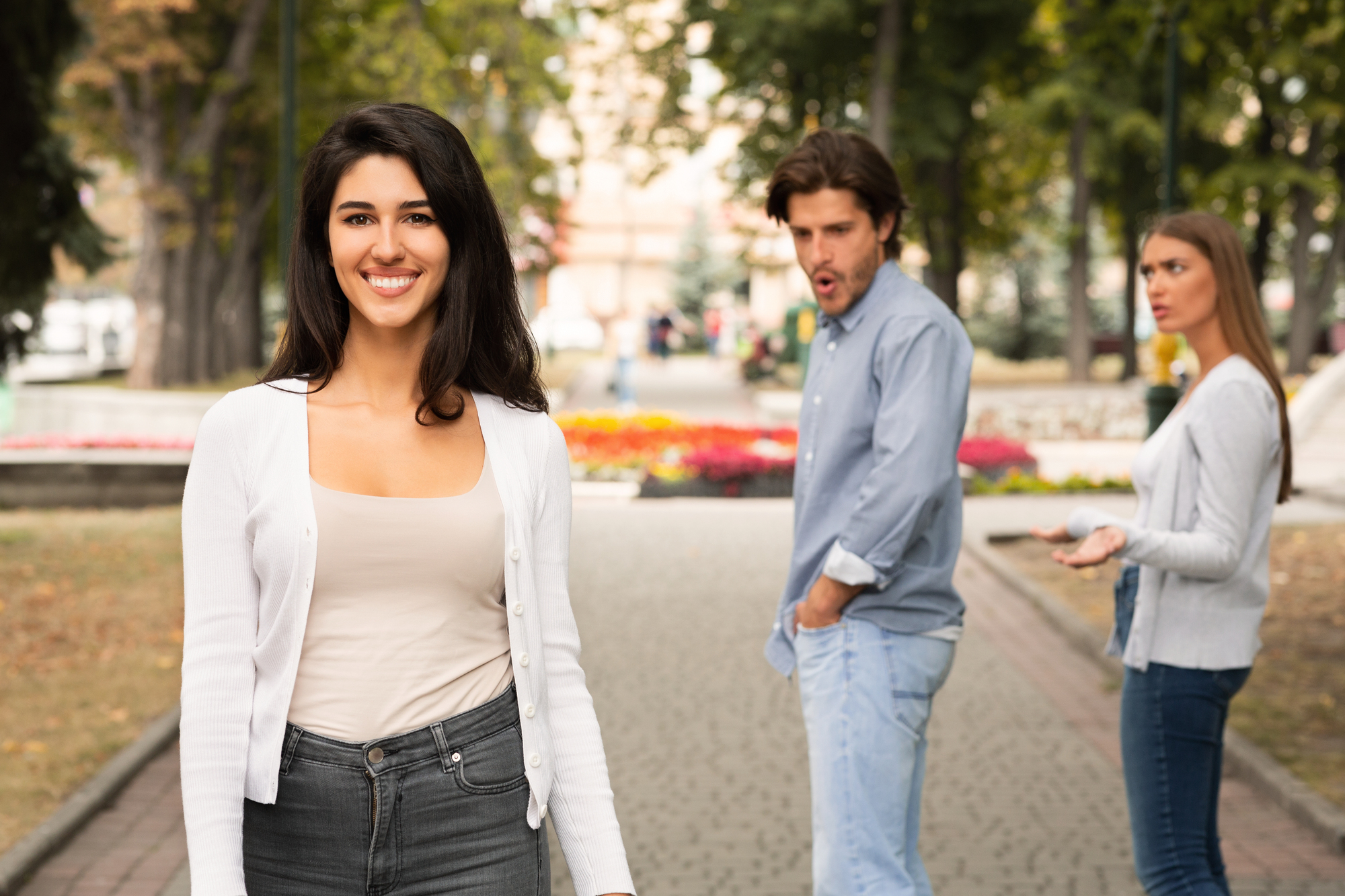 A woman smiling in the foreground as a man looks back at her with surprise, while a woman with him looks annoyed. They are on a paved path in a park, surrounded by trees and colorful flowers.