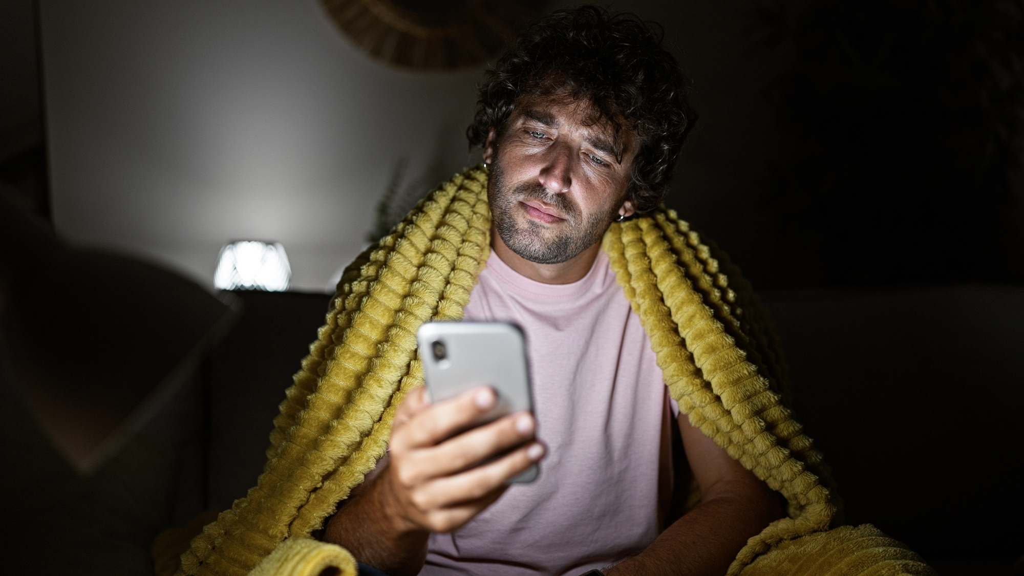 A man sitting on a couch, wrapped in a yellow blanket, looks intently at his smartphone screen. The room is dimly lit, with a small lamp in the background providing soft lighting. He wears a light-pink shirt.