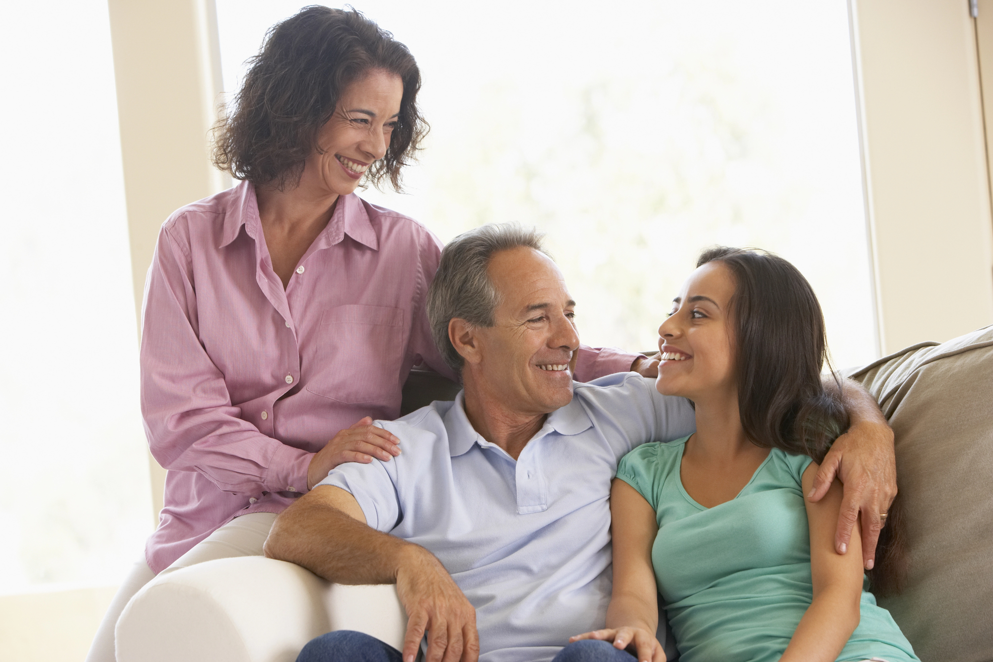 A family of three sits together on a sofa, smiling and talking. The woman on the left stands behind the man in the middle, resting her hand on his shoulder, while the young woman on the right sits beside him. Bright light filters through the windows.