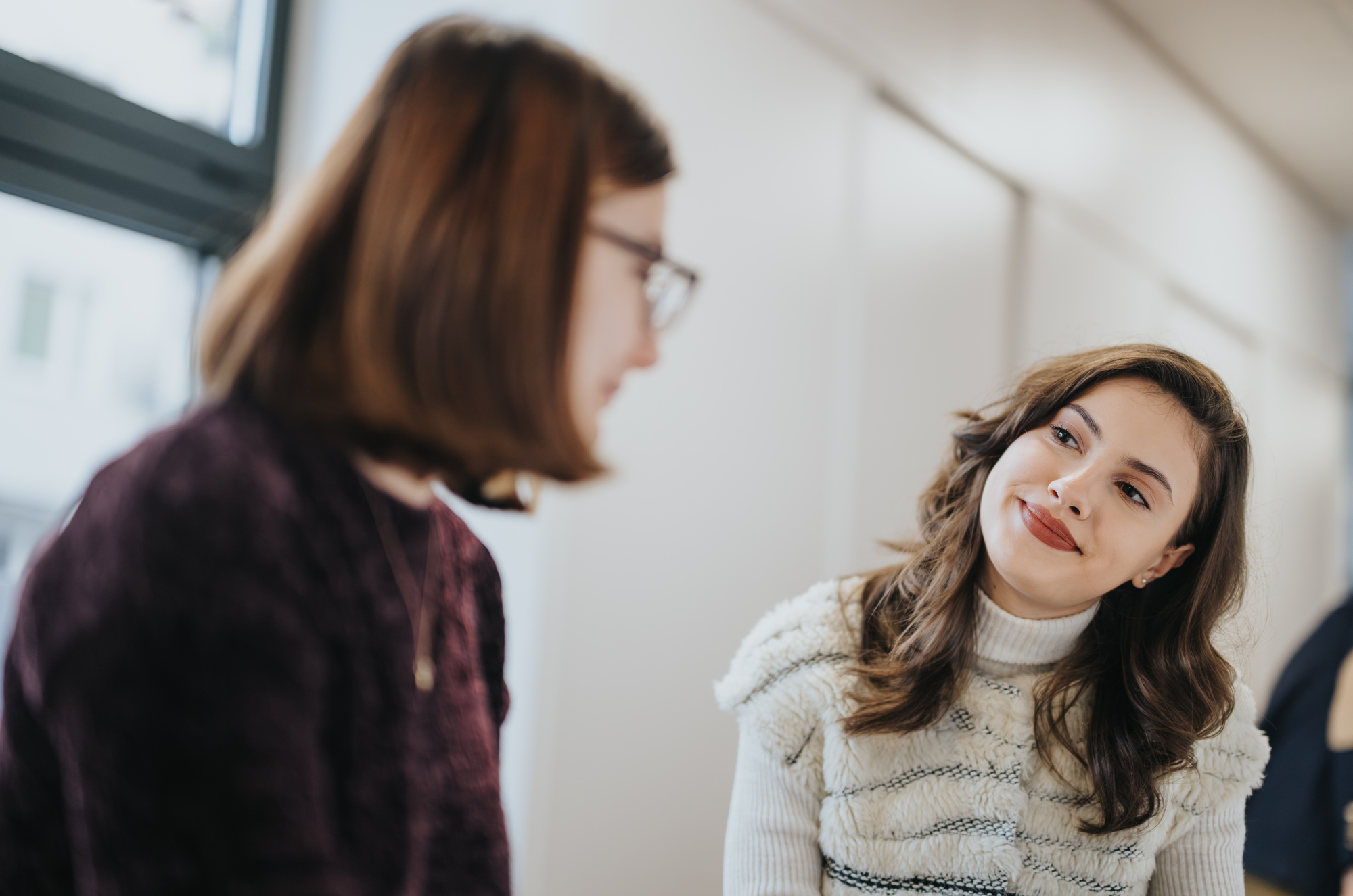 Two women are sitting indoors, engaged in conversation. One woman, with long brown hair, is smiling and listening intently to the other woman, who is speaking. The setting appears casual and well-lit by natural light.