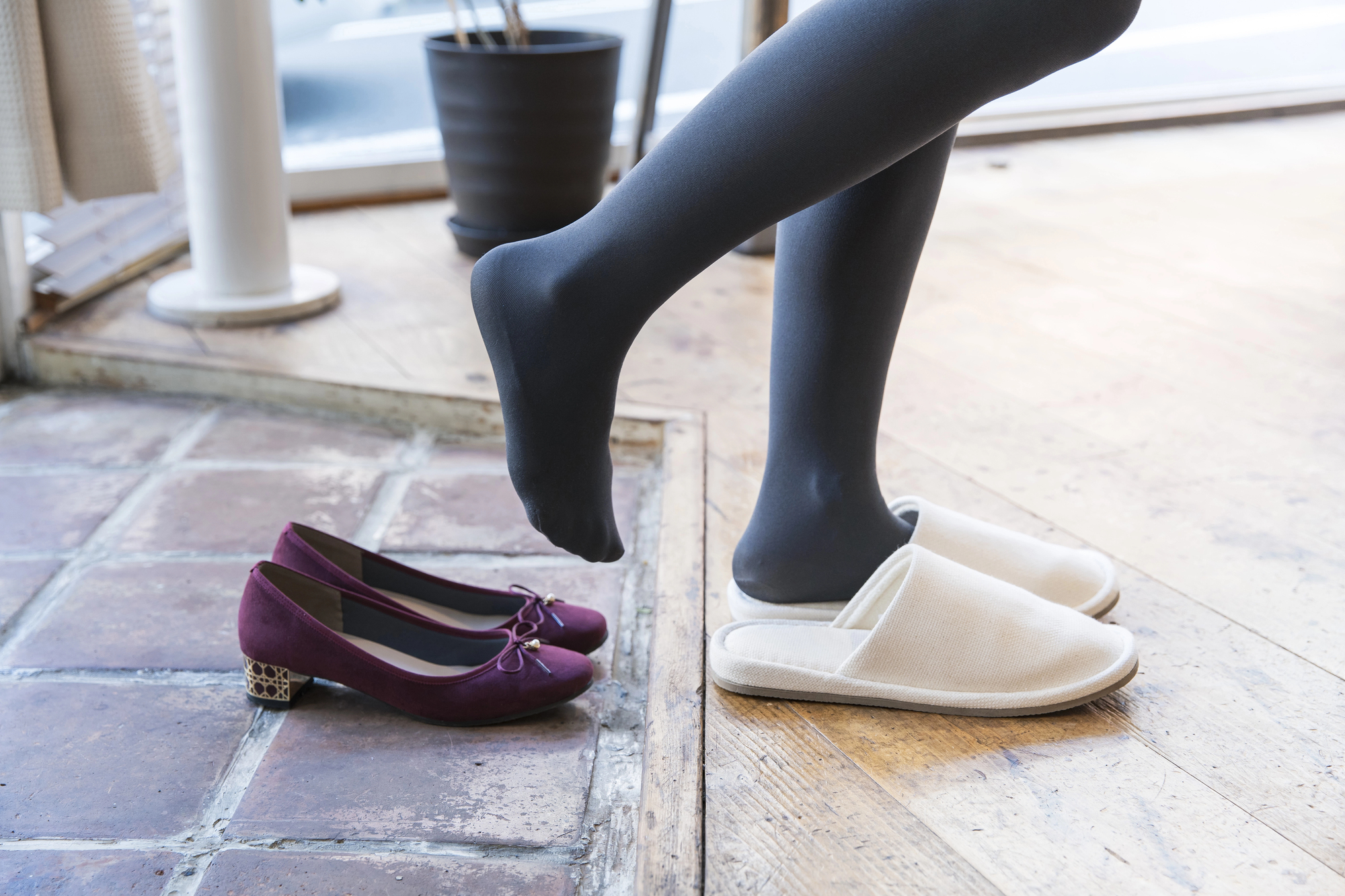 A person wearing black tights steps into white slippers beside a pair of purple shoes with decorative heels on a wooden floor. A potted plant and a window are in the background.