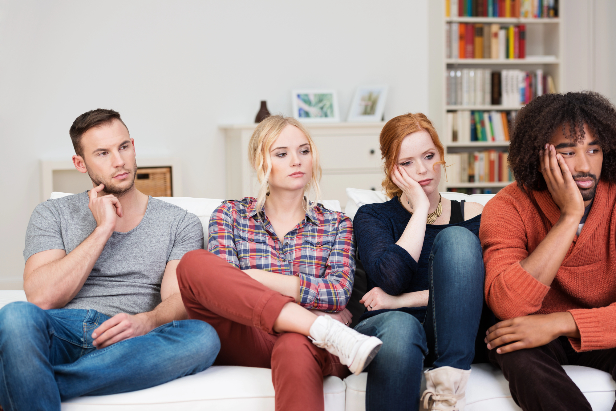 Four people sitting on a couch, all appearing bored or uninterested. They rest their heads on their hands, gazing off into the distance. The room has a bookshelf and various decorative elements in the background.