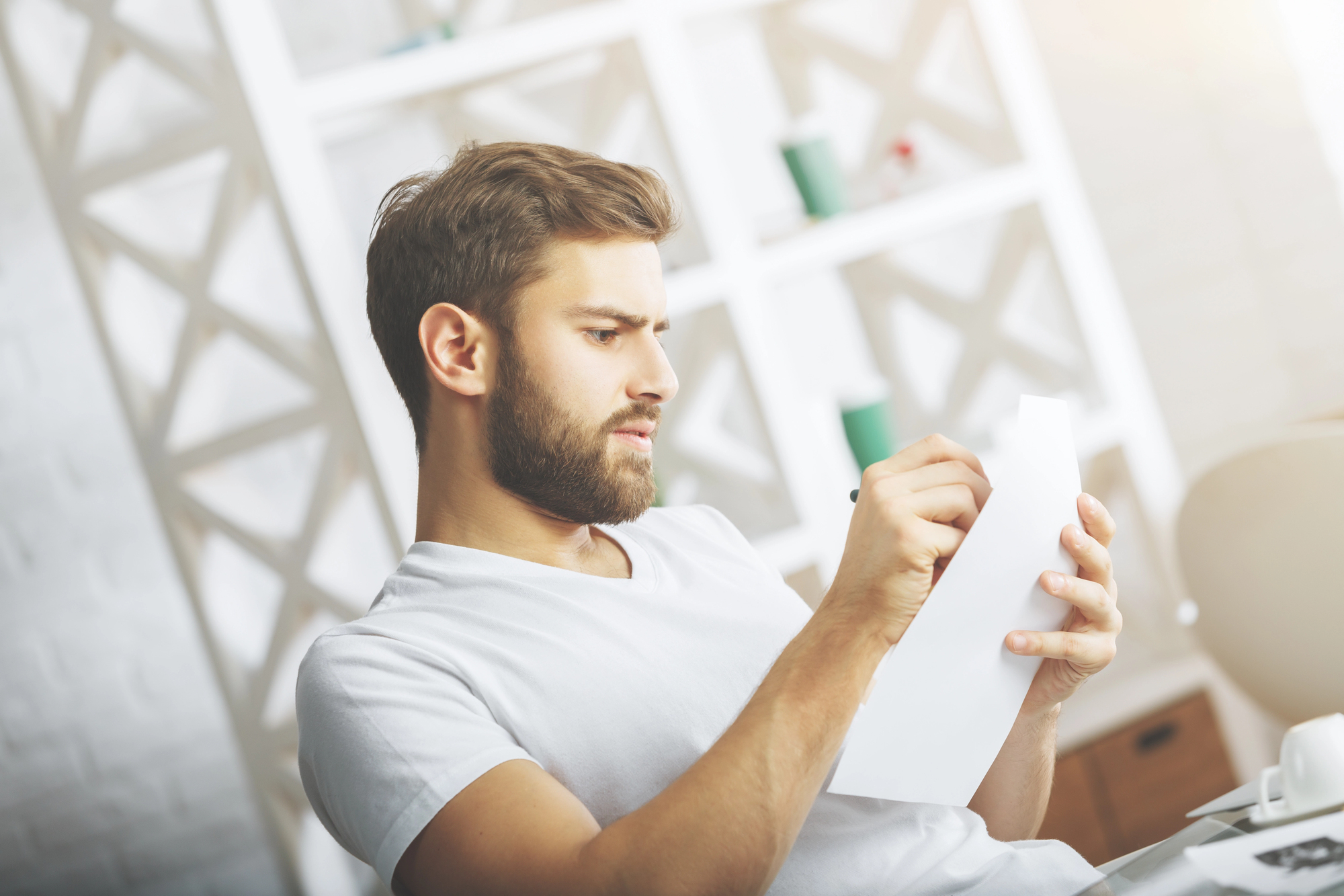 A man with a beard wearing a white T-shirt is sitting in a modern office environment, intently reading and writing on a piece of paper. There are shelves with decor items in the background, and soft sunlight illuminates the scene.