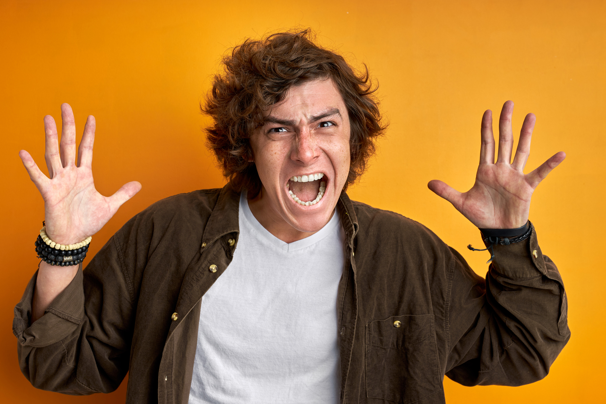 A person with curly hair and a white t-shirt under a brown jacket stands against an orange background, raising both hands with splayed fingers and an open-mouthed, expressive face.