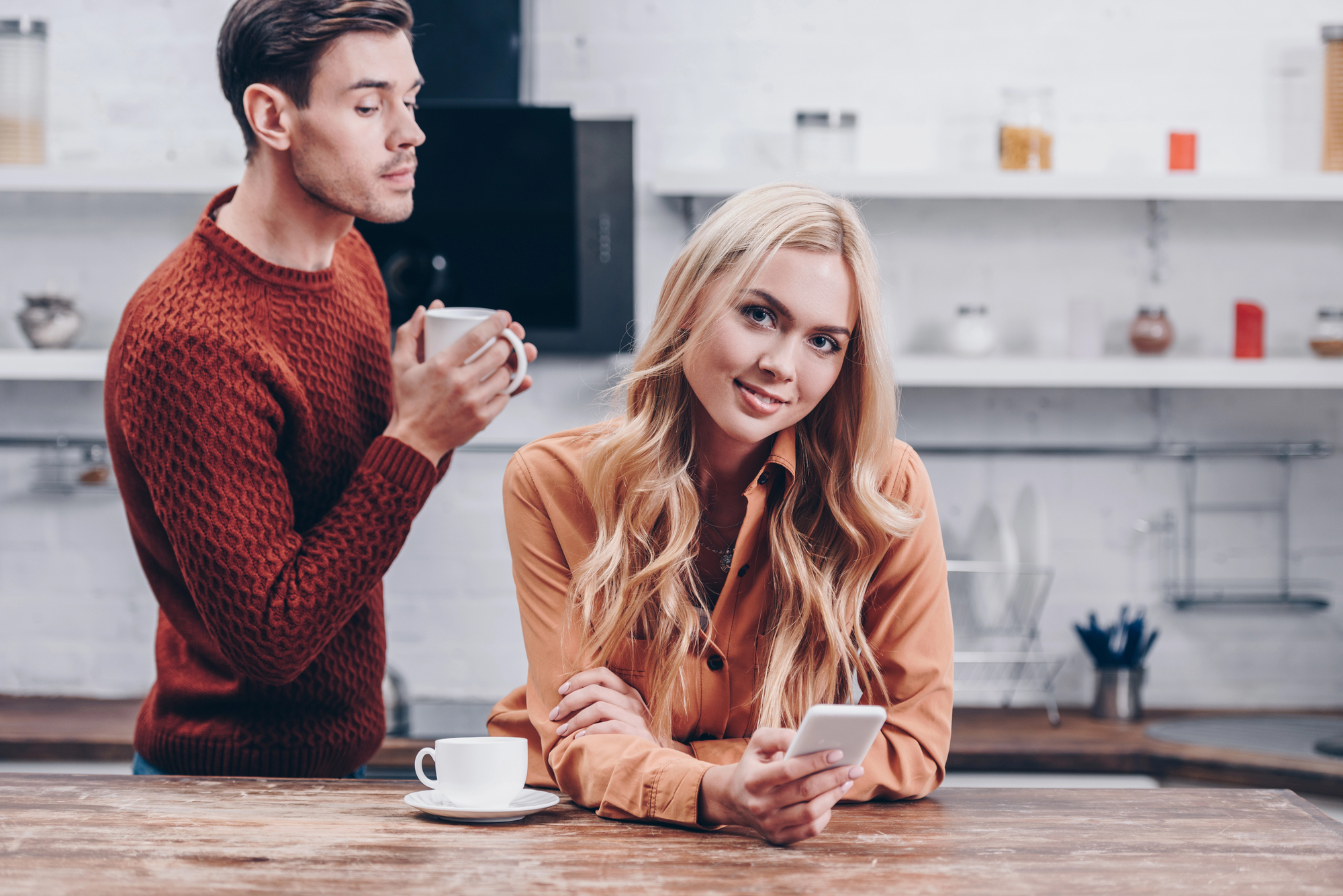 A woman with long blonde hair uses a smartphone, smiling slightly, while sitting at a wooden counter. A man with short hair, wearing a red sweater, stands nearby holding a white mug. The background features white brick walls and shelves.