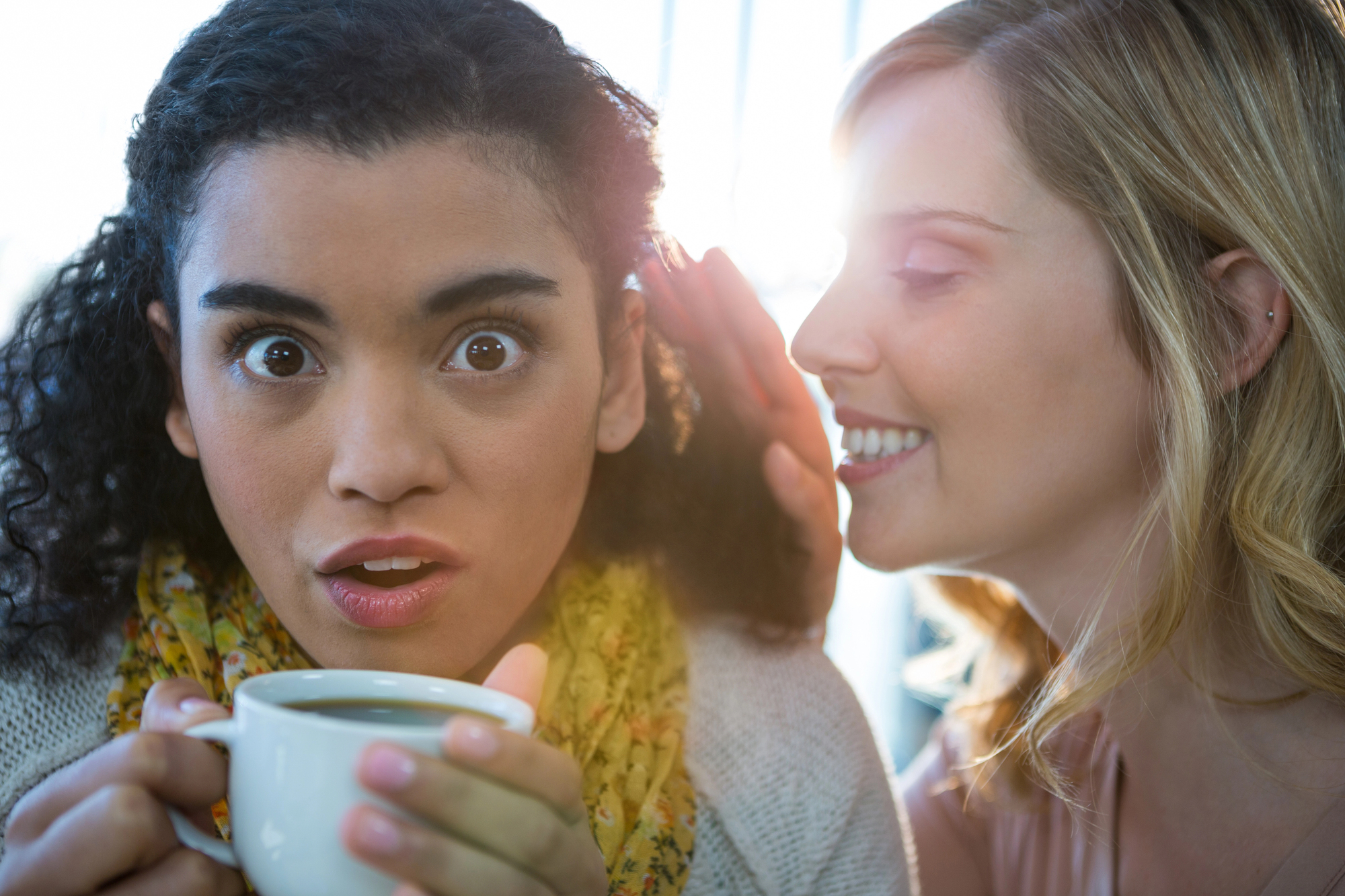 A surprised woman is holding a white mug while another woman whispers in her ear with a smile. Sunlight streams through the window behind them, creating a bright, warm atmosphere.