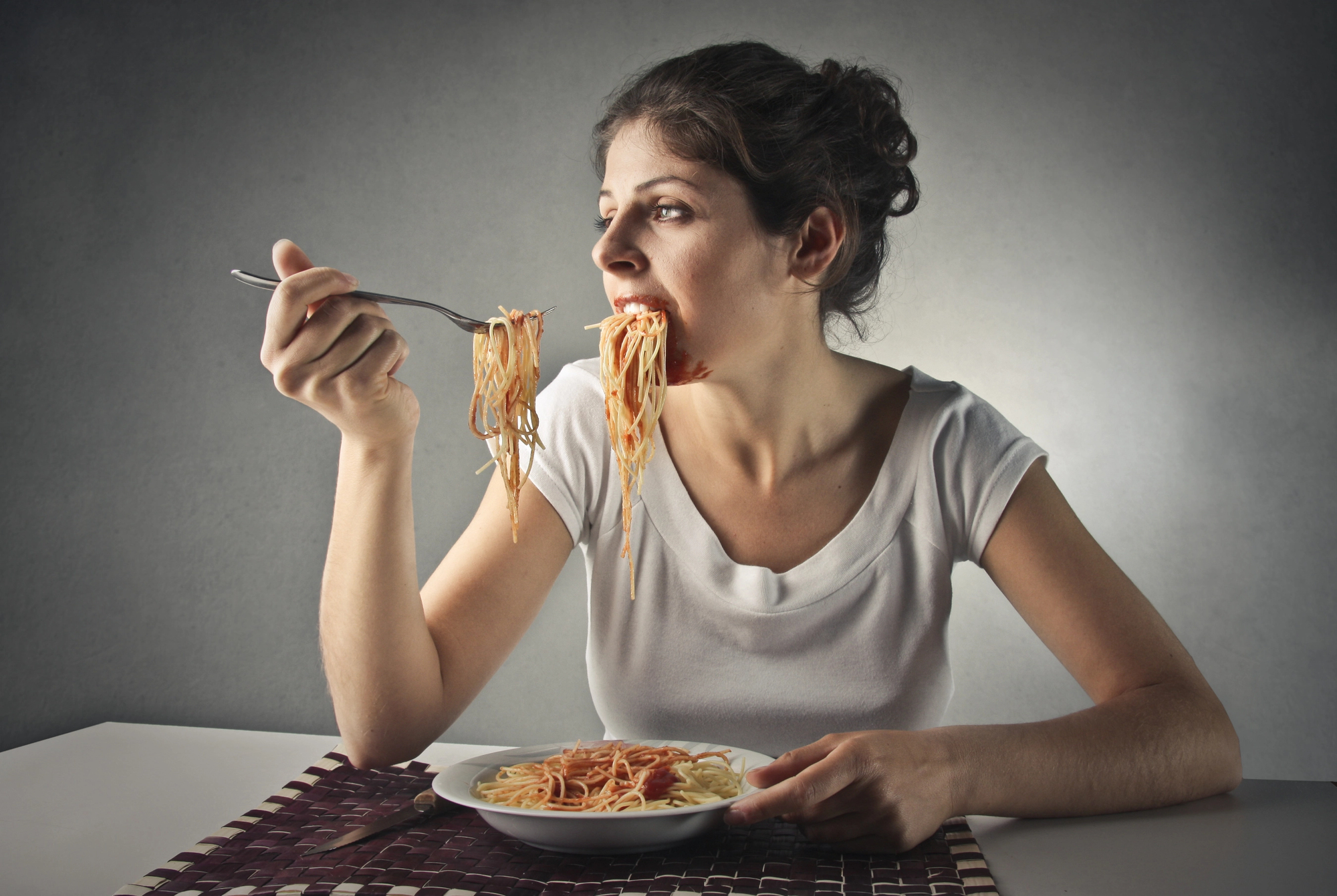 A woman in a white shirt is sitting at a table eating spaghetti with a fork. She looks focused on her meal, with a plate of pasta in front of her and a woven placemat on the table. The background is a plain gray wall.