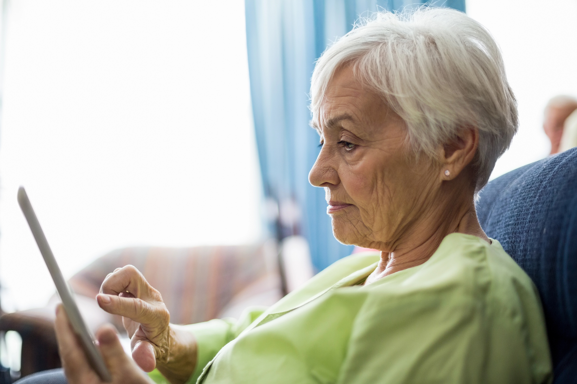 An older woman with short white hair sits on a sofa, focused on a tablet in her hand. She wears a light green top, and the room around her is softly lit with a blurred background.