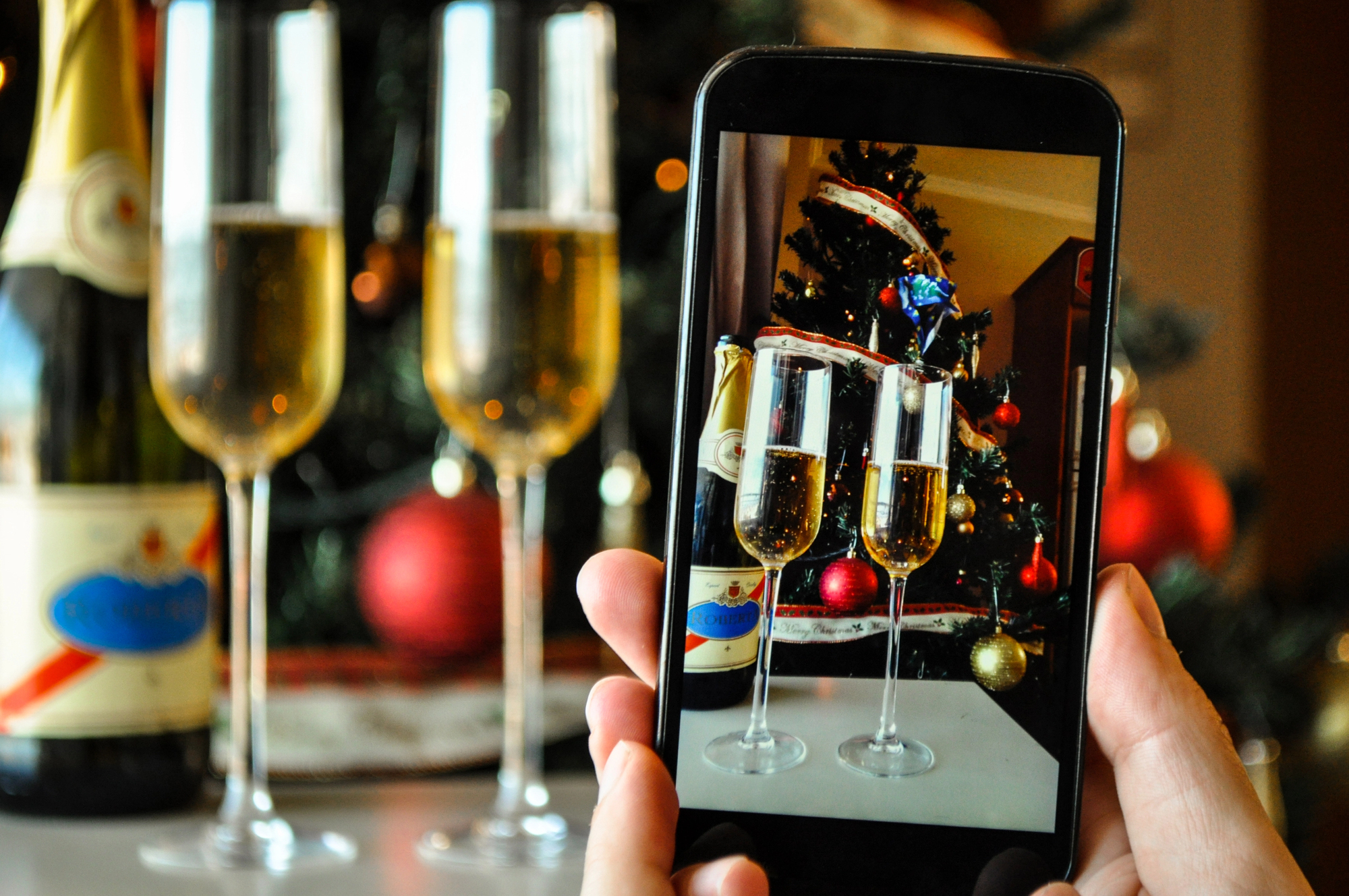 A person takes a photo of three champagne glasses with a smartphone. The background shows a decorated Christmas tree and a champagne bottle. The focus is on the phone screen displaying the glasses.