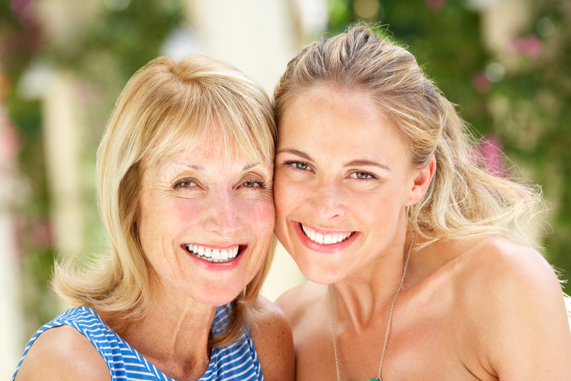 A smiling older woman with short blonde hair and a younger woman with long blonde hair pose closely together outdoors, with greenery in the background. They both wear sleeveless tops and express a joyful mood.