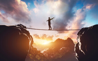 A person balances on a tightrope stretched between two cliffs at sunset. The sky is filled with dramatic clouds, and the sun is partially visible on the horizon, casting a warm glow over the mountainous landscape.