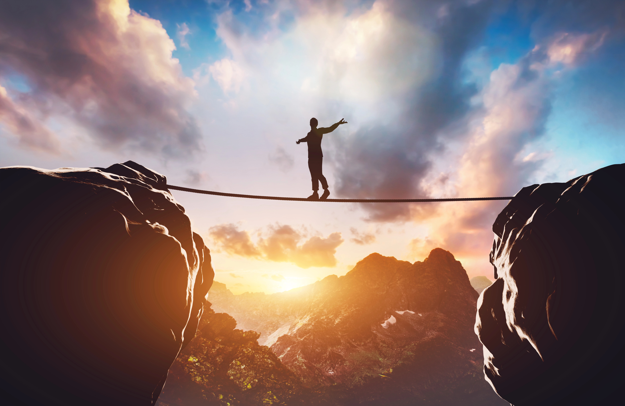 A person balances on a tightrope stretched between two cliffs at sunset. The sky is filled with dramatic clouds, and the sun is partially visible on the horizon, casting a warm glow over the mountainous landscape.