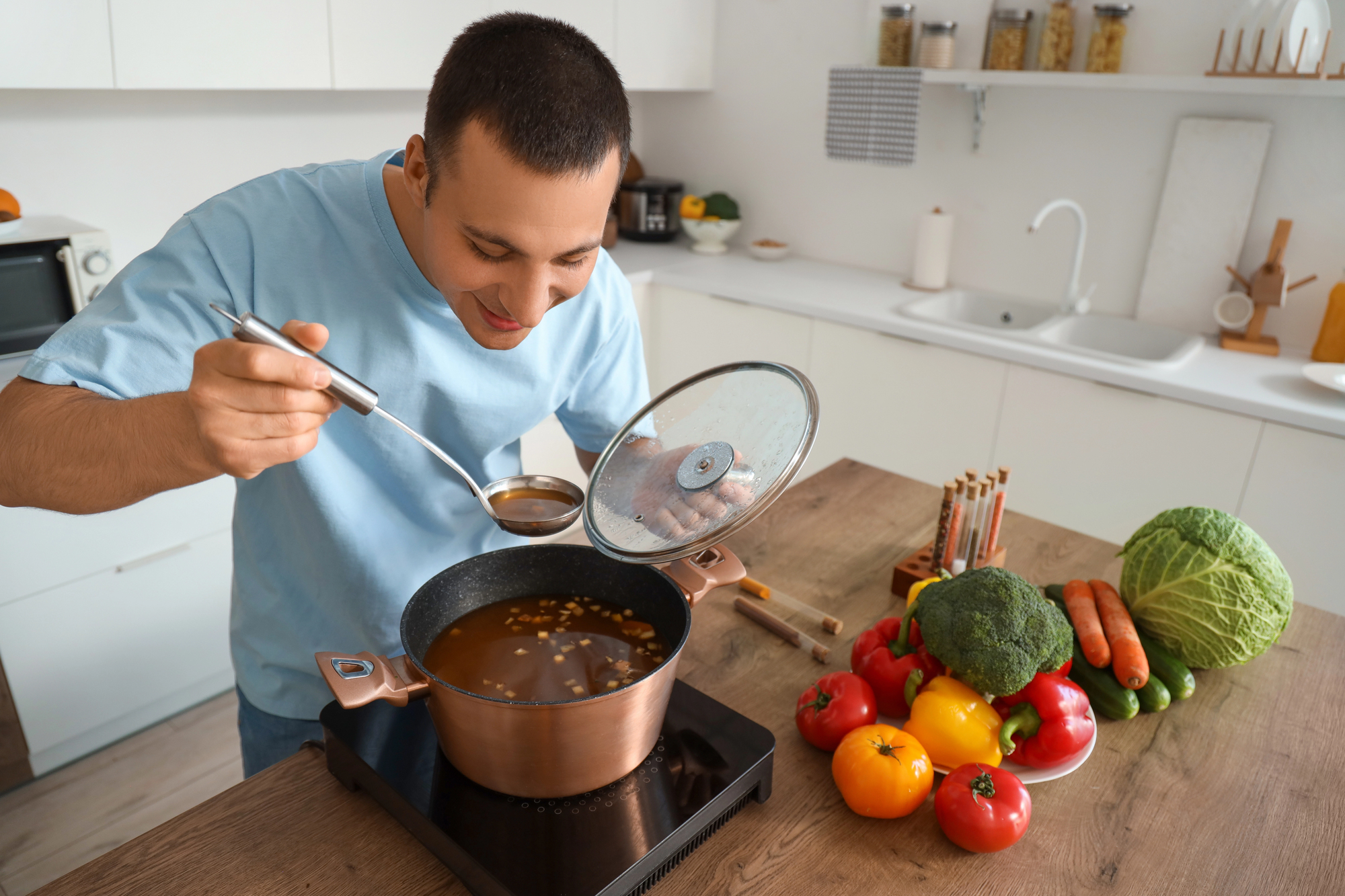 A man in a light blue shirt tastes soup from a copper pot on a stove in a modern kitchen. Colorful vegetables like tomatoes, broccoli, carrots, and cabbage are on the wooden countertop beside him.