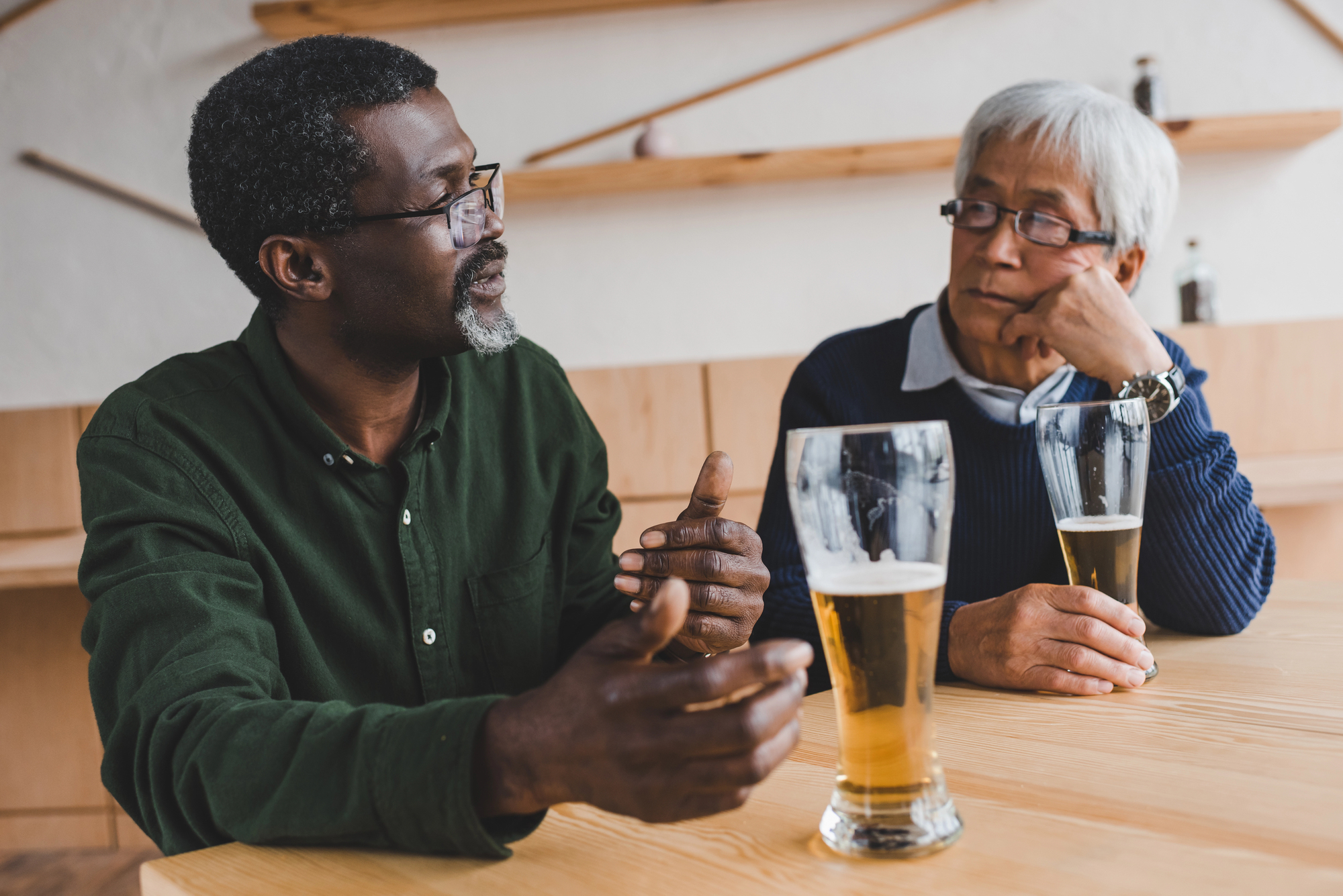 Two men are seated at a wooden table, each holding a glass of beer. The man on the left is gesturing while speaking, and the man on the right is listening intently, resting his chin on his hand. They are in a modern, minimalist setting.