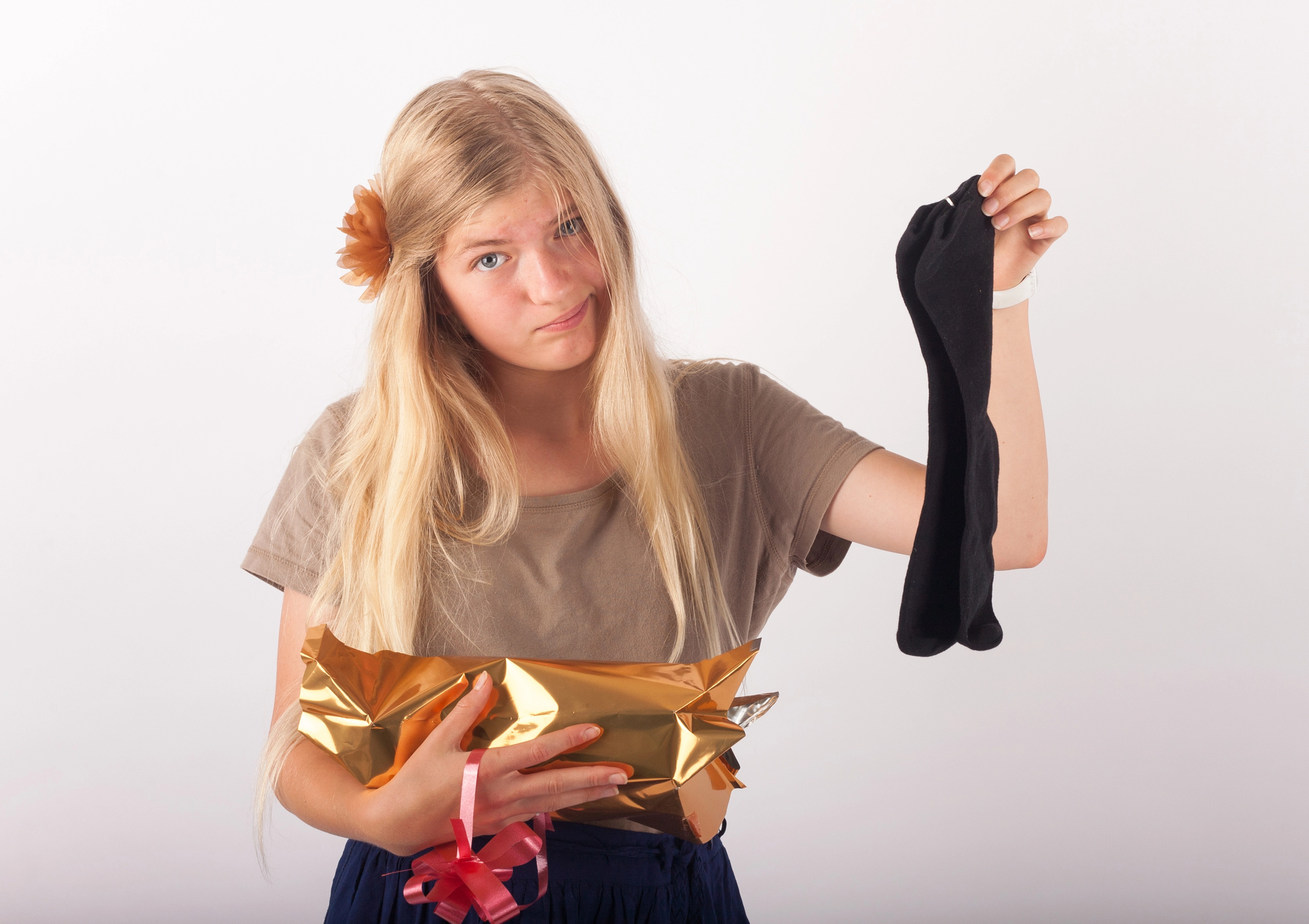 A young girl with long blonde hair holds up a black sock and a gold-wrapped gift, looking disappointed. She is wearing a brown top and has a flower hair accessory. The background is plain white.