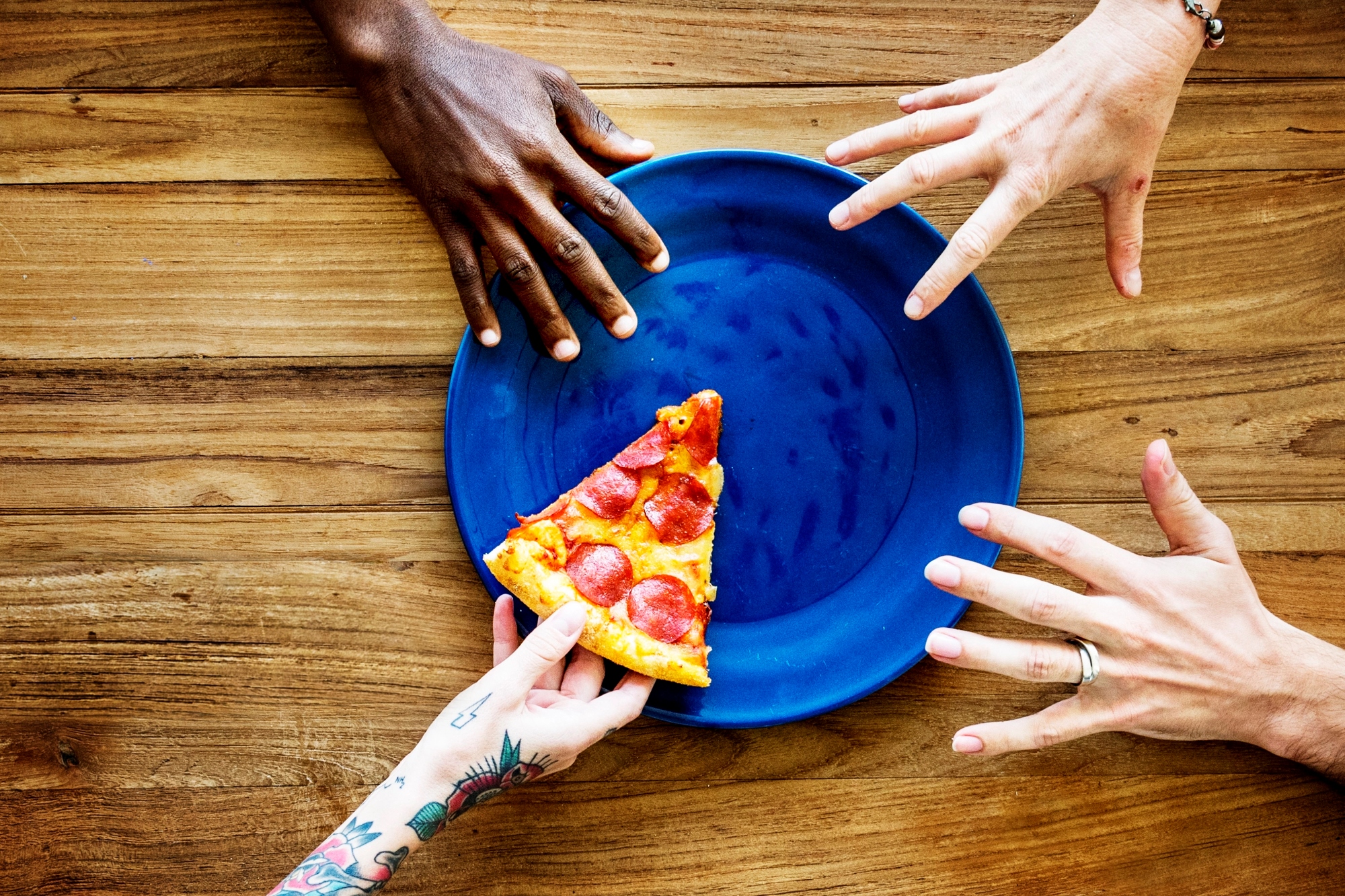 Four hands reaching toward a blue plate on a wooden table, with one hand holding a slice of pepperoni pizza.