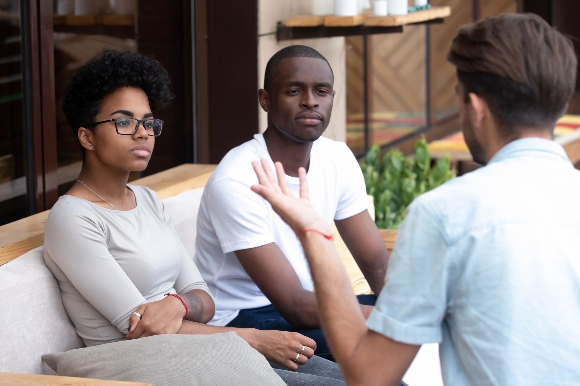 Three people are sitting on a couch having a discussion. The two facing the camera look attentive, while the person with their back to the camera gestures with their hand. They are in an indoor setting with plants in the background.