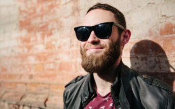 A bearded man wearing sunglasses and a black leather jacket stands against a sunlit brick wall, smiling contentedly. Shadows are cast on the wall, and the man appears to be enjoying a sunny day.