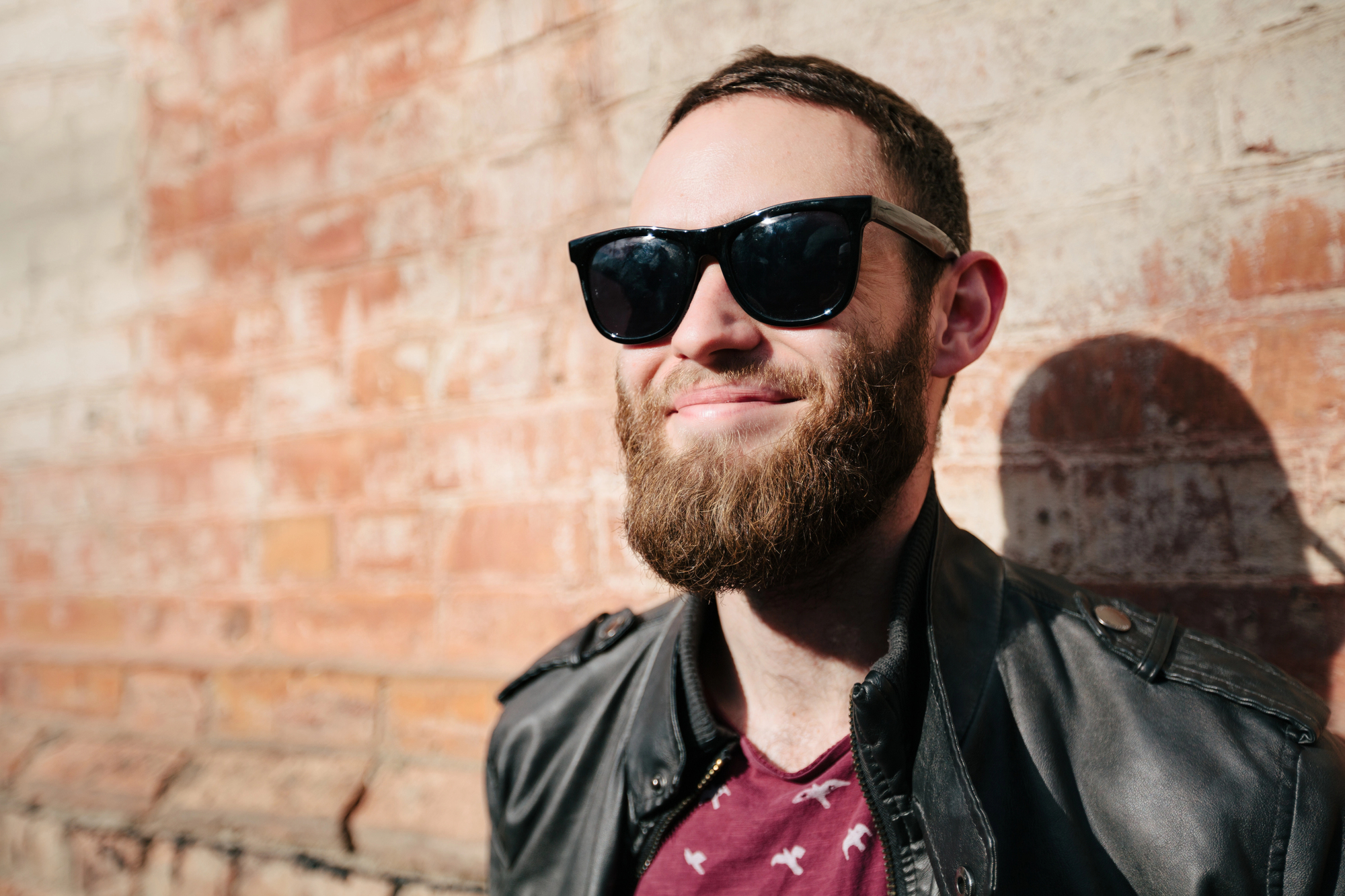 A bearded man wearing sunglasses and a black leather jacket stands against a sunlit brick wall, smiling contentedly. Shadows are cast on the wall, and the man appears to be enjoying a sunny day.