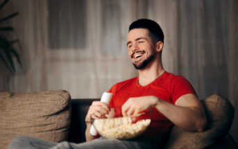 A man in a red shirt sits on a couch, holding a remote control and a bowl of popcorn. He is smiling and appears to be enjoying a show or movie. The background is softly lit, creating a cozy atmosphere.