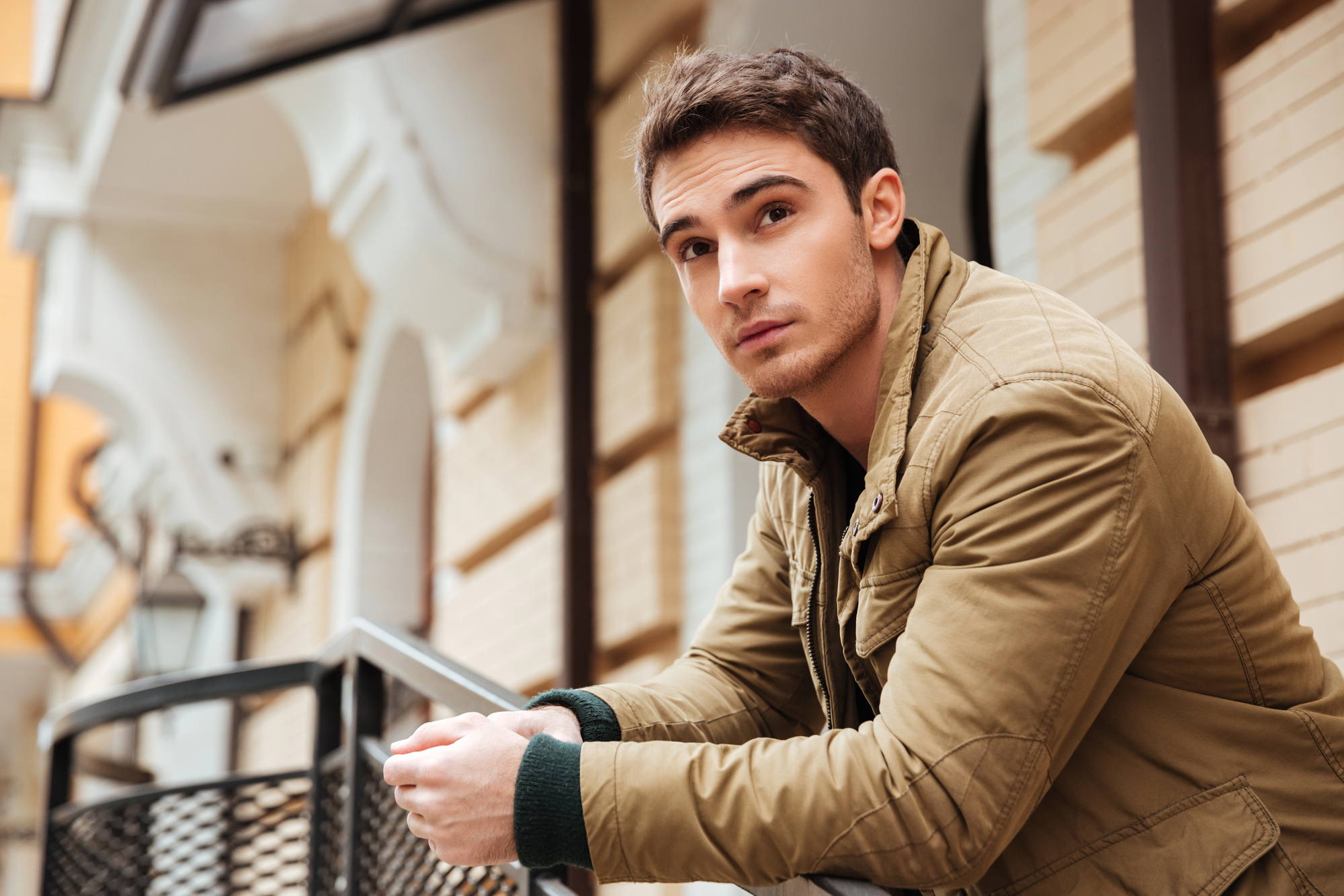 A young man with short brown hair, wearing a brown jacket, leans on a railing outdoors. The background shows a brick building and a street lamp. He gazes thoughtfully into the distance.