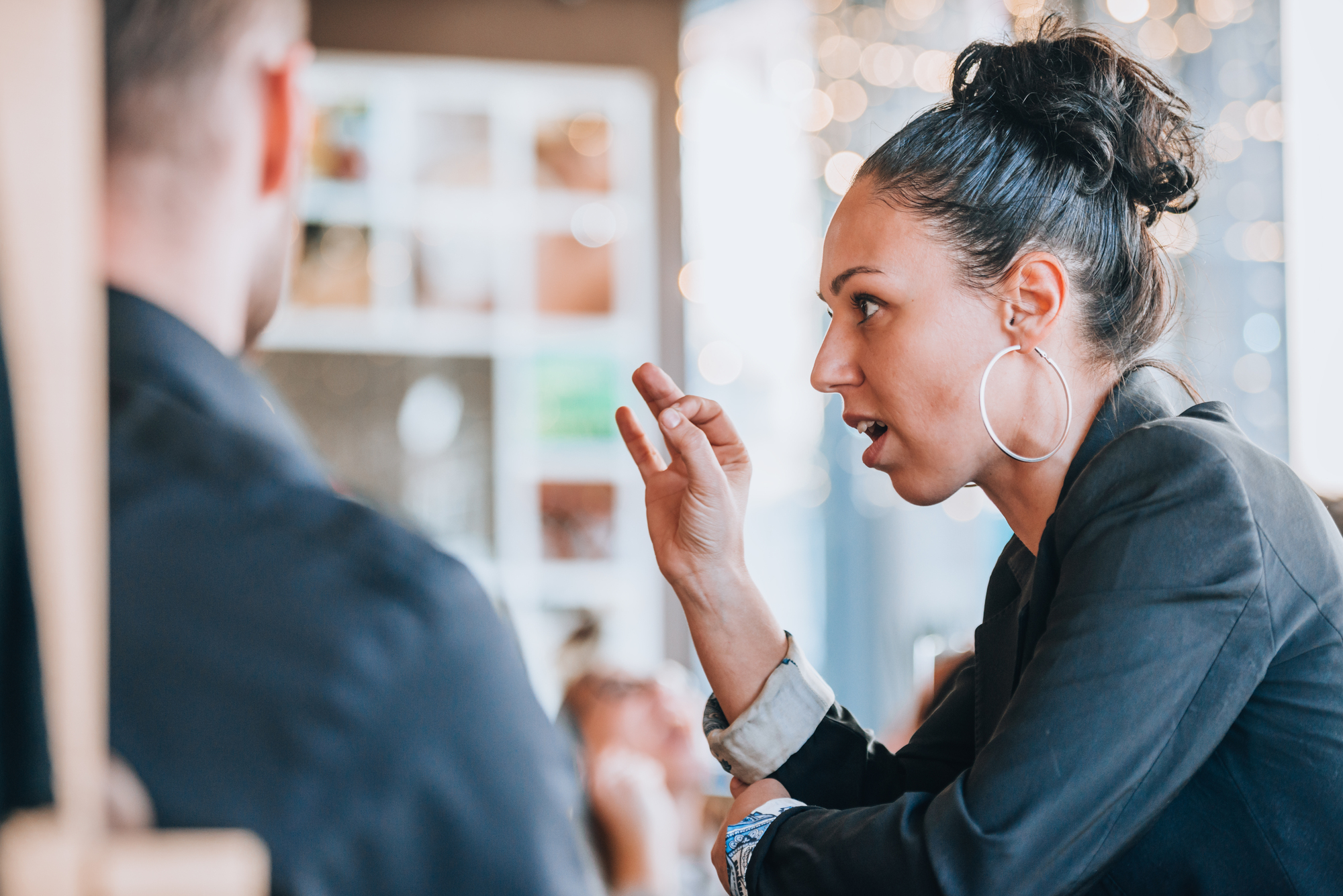 Woman with hoop earrings conversing with a man in a dimly lit cafe. She gestures with her hand, appearing engaged in the discussion. The background is softly blurred with decorative lights.