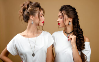 Two women with similar hairstyles and makeup stand facing each other against a beige background. Both wear white tops and heart-shaped necklaces, with one having a braid and the other a bun. Their lips are painted red.