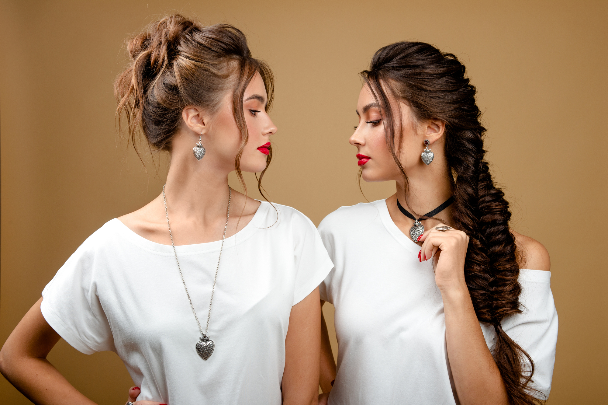 Two women with similar hairstyles and makeup stand facing each other against a beige background. Both wear white tops and heart-shaped necklaces, with one having a braid and the other a bun. Their lips are painted red.