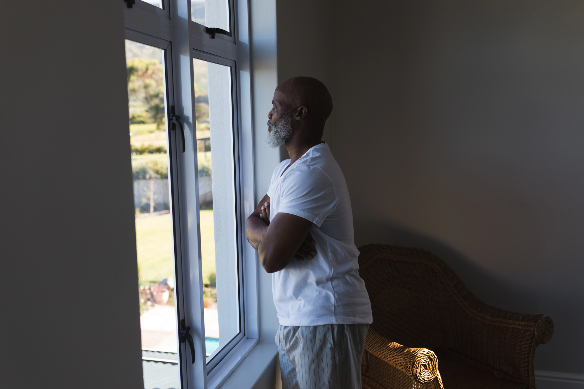 A man with a beard, wearing a white t-shirt, stands indoors looking out of a large window. Sunlight illuminates the room, highlighting a wicker chair nearby. Outside, greenery and a clear sky are visible.