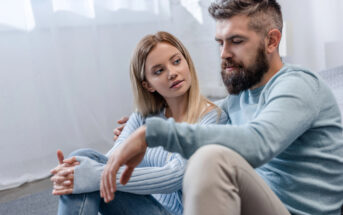 A woman and a man sit together on the floor in a brightly lit room, both wearing light blue tops and light-colored pants. The woman looks intently at the man as he speaks, creating a sense of a serious or meaningful conversation.