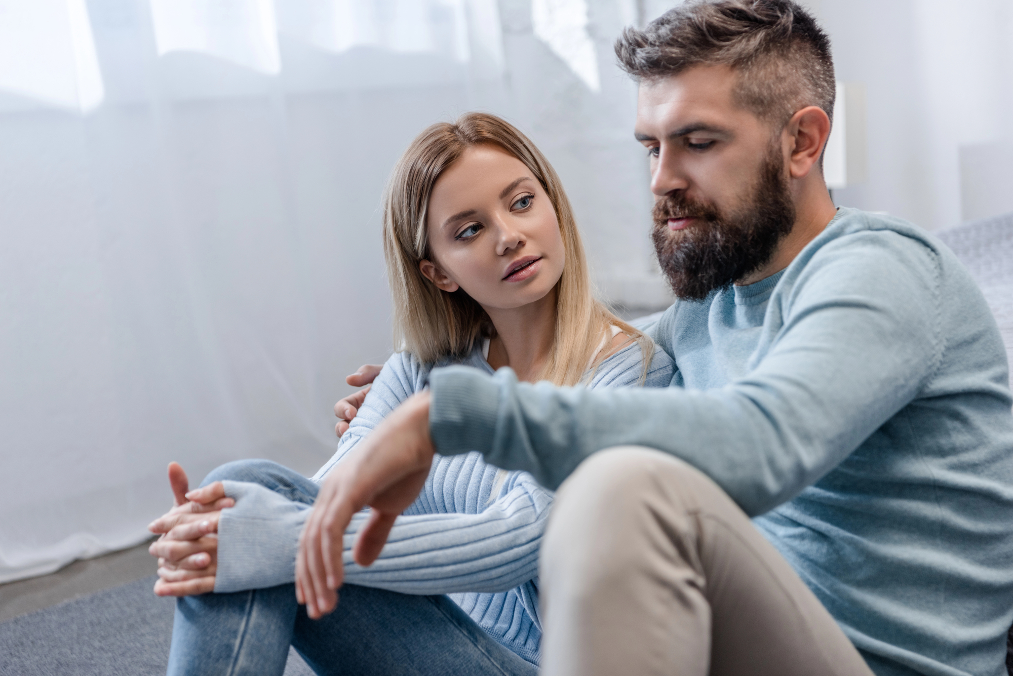 A woman and a man sit together on the floor in a brightly lit room, both wearing light blue tops and light-colored pants. The woman looks intently at the man as he speaks, creating a sense of a serious or meaningful conversation.