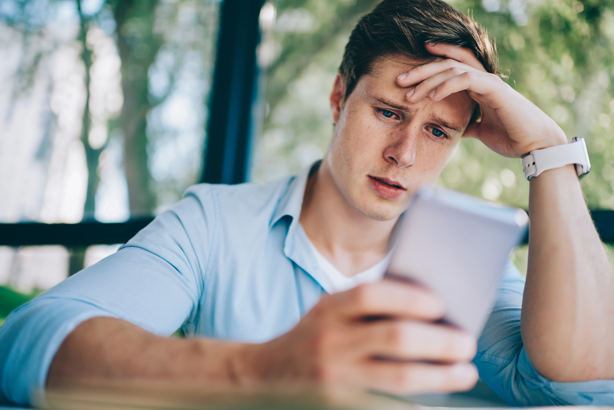 A young man looks at his smartphone with a concerned expression, holding his forehead with one hand. He is wearing a light blue shirt and a white watch. Blurred greenery is visible in the background.