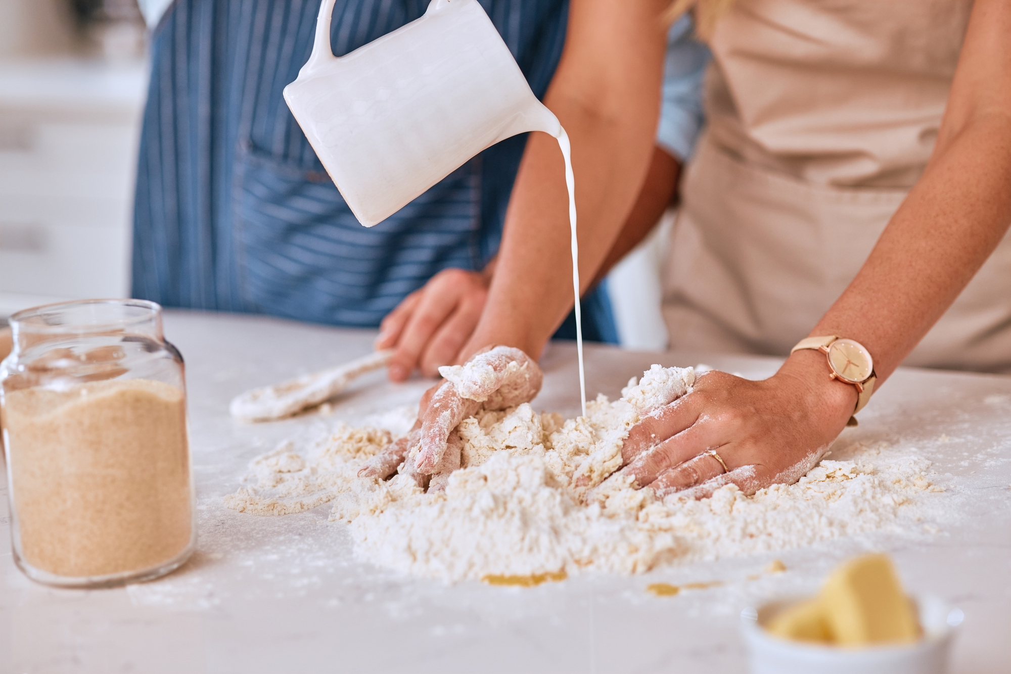 Two people baking together. One person pours milk from a pitcher into a pile of flour on a countertop while the other mixes the ingredients with their hands. A jar of sugar and some butter are visible nearby. Both are wearing aprons.