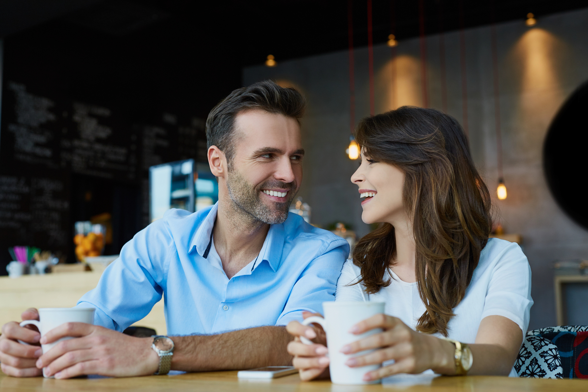 A man and a woman sit at a café table, smiling at each other while holding white mugs. The background features a menu board and hanging lights, creating a cozy atmosphere.