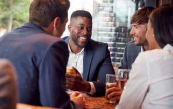 Four people in business attire sit around a table in a cafe, engaged in a lively conversation. One man, holding a drink, appears to be speaking, while the others listen attentively with smiles. The atmosphere is warm and casual.