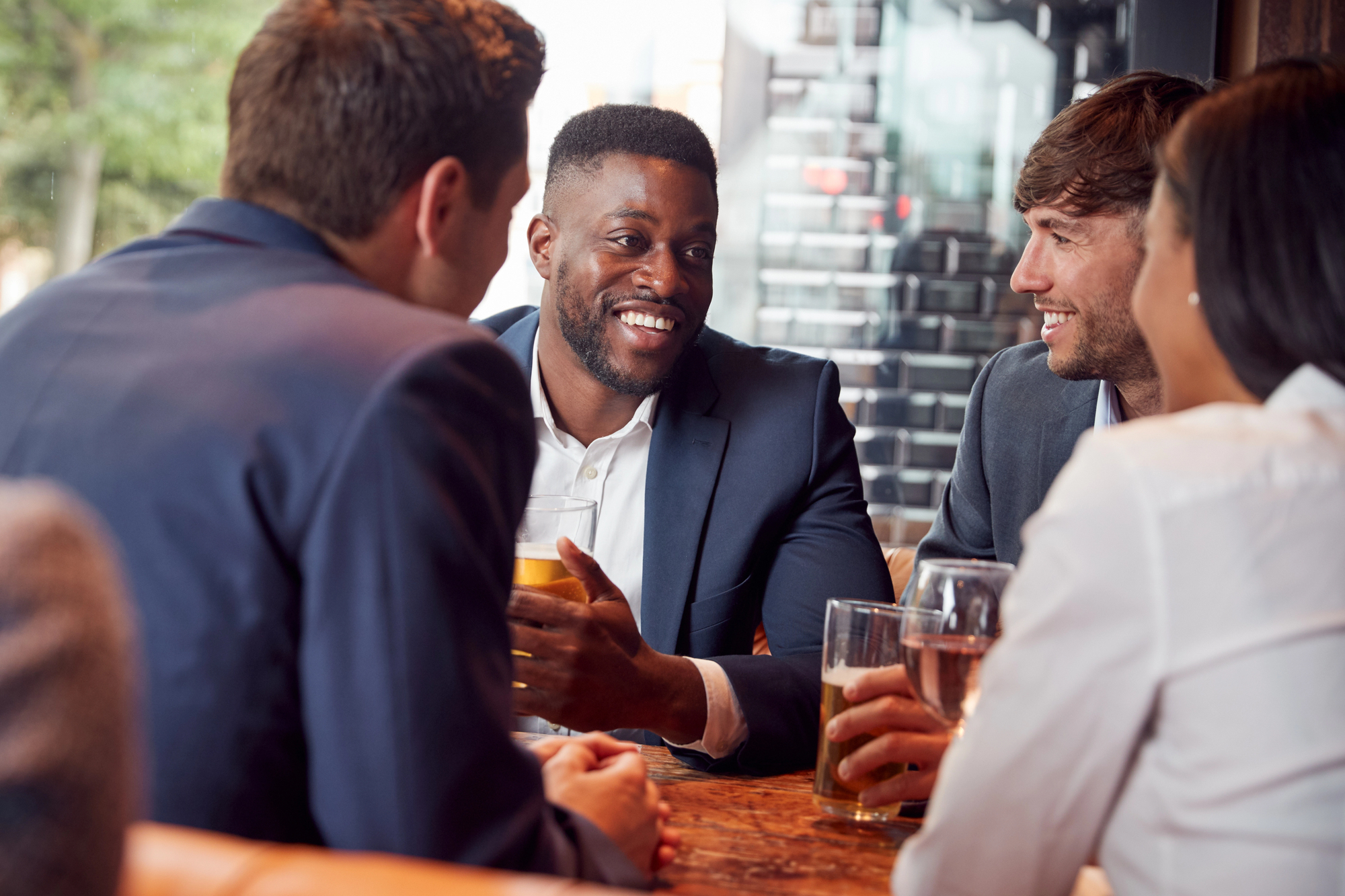 Four people in business attire sit around a table in a cafe, engaged in a lively conversation. One man, holding a drink, appears to be speaking, while the others listen attentively with smiles. The atmosphere is warm and casual.