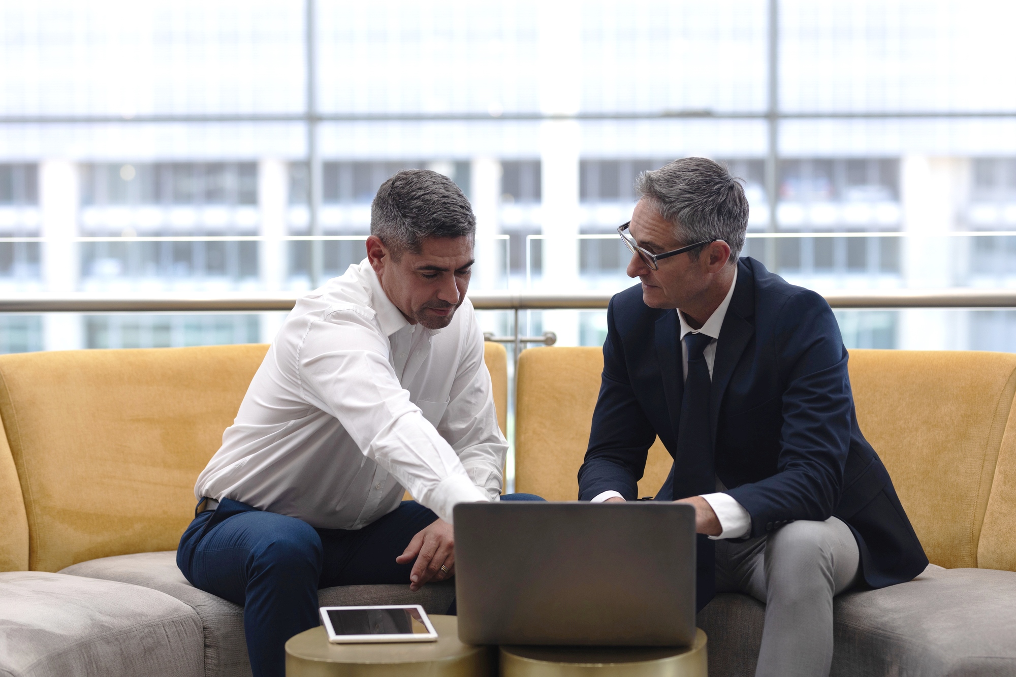 Two men sit on a yellow couch in a modern office setting, engaged in conversation while looking at a laptop in front of them. One man points at the screen as they discuss. A tablet lies on the table beside them.