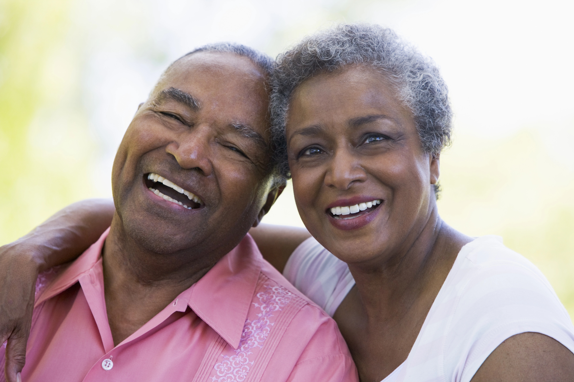 An older couple is smiling and embracing outdoors. The man is wearing a pink shirt, and the woman is wearing a white blouse. The background is blurred with green and yellow tones, suggesting a natural setting.