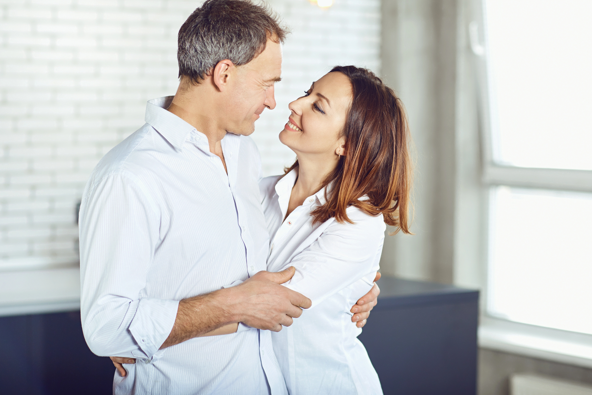 A couple in white shirts embraces and smiles at each other in a bright room with a white brick wall and large window in the background.