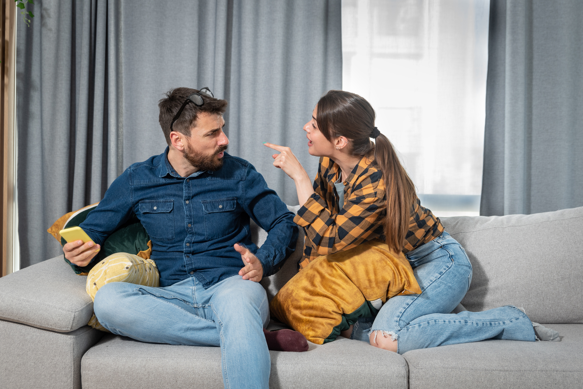 A man and a woman sit on a couch, engaged in an argument. The man in a denim shirt looks frustrated, holding a phone. The woman in a plaid shirt points her finger at him, expressing anger. Grey curtains and cushions form the background.