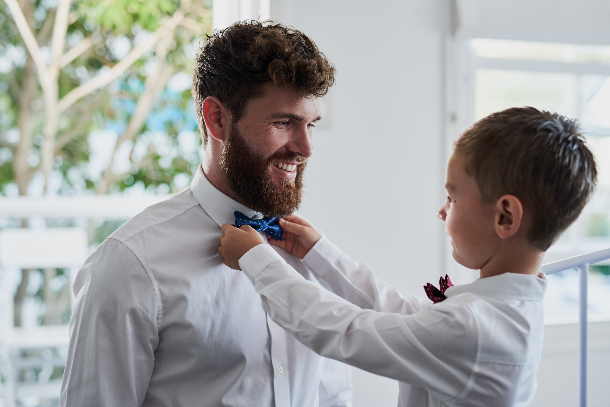 A young boy adjusts a bow tie for a smiling man with a beard. Both are wearing white shirts. They are indoors, with a window and greenery visible in the background.