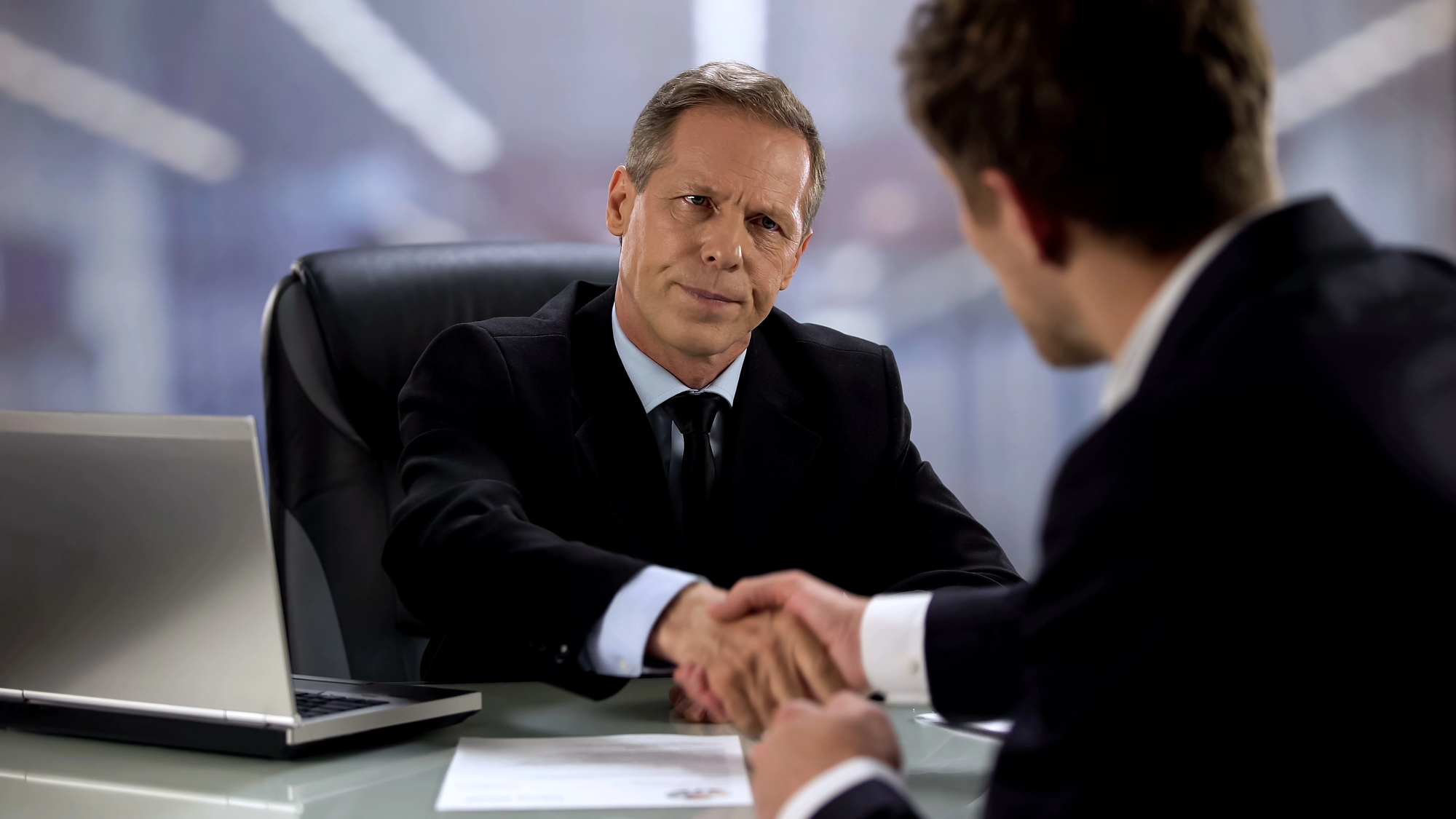 Two men in suits shaking hands across a desk in an office setting. One man faces the camera with a serious expression, while the other man's back is to the camera. A laptop and documents are on the desk.