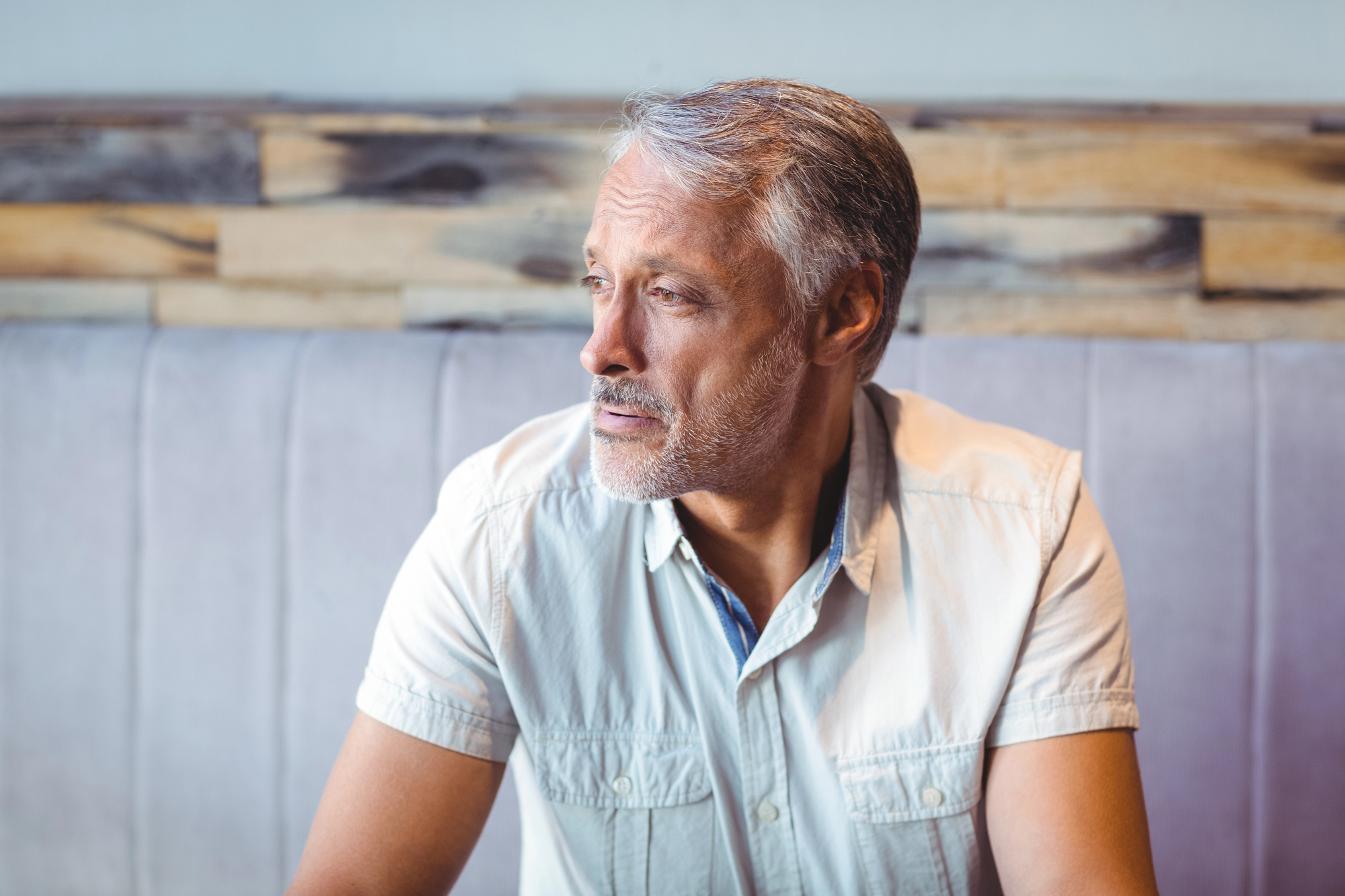 A middle-aged man with gray hair and facial stubble gazes to the side, seated in a booth with a wooden panel backdrop. He wears a short-sleeved light gray shirt and appears thoughtful.