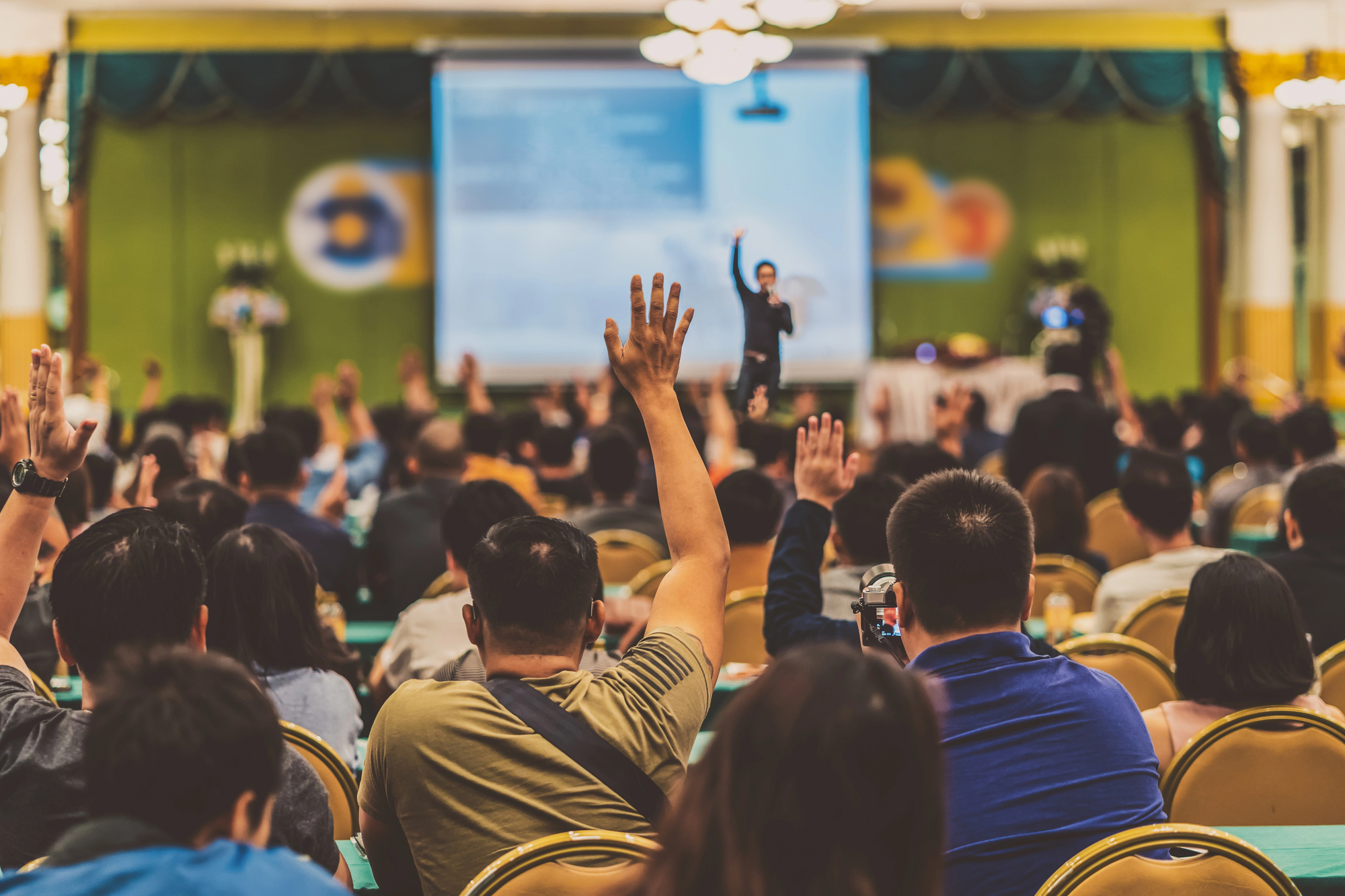 A speaker stands in front of a large audience in a conference room, with participants raising their hands. The focus is on the audience, while a blurred presentation screen is visible in the background.