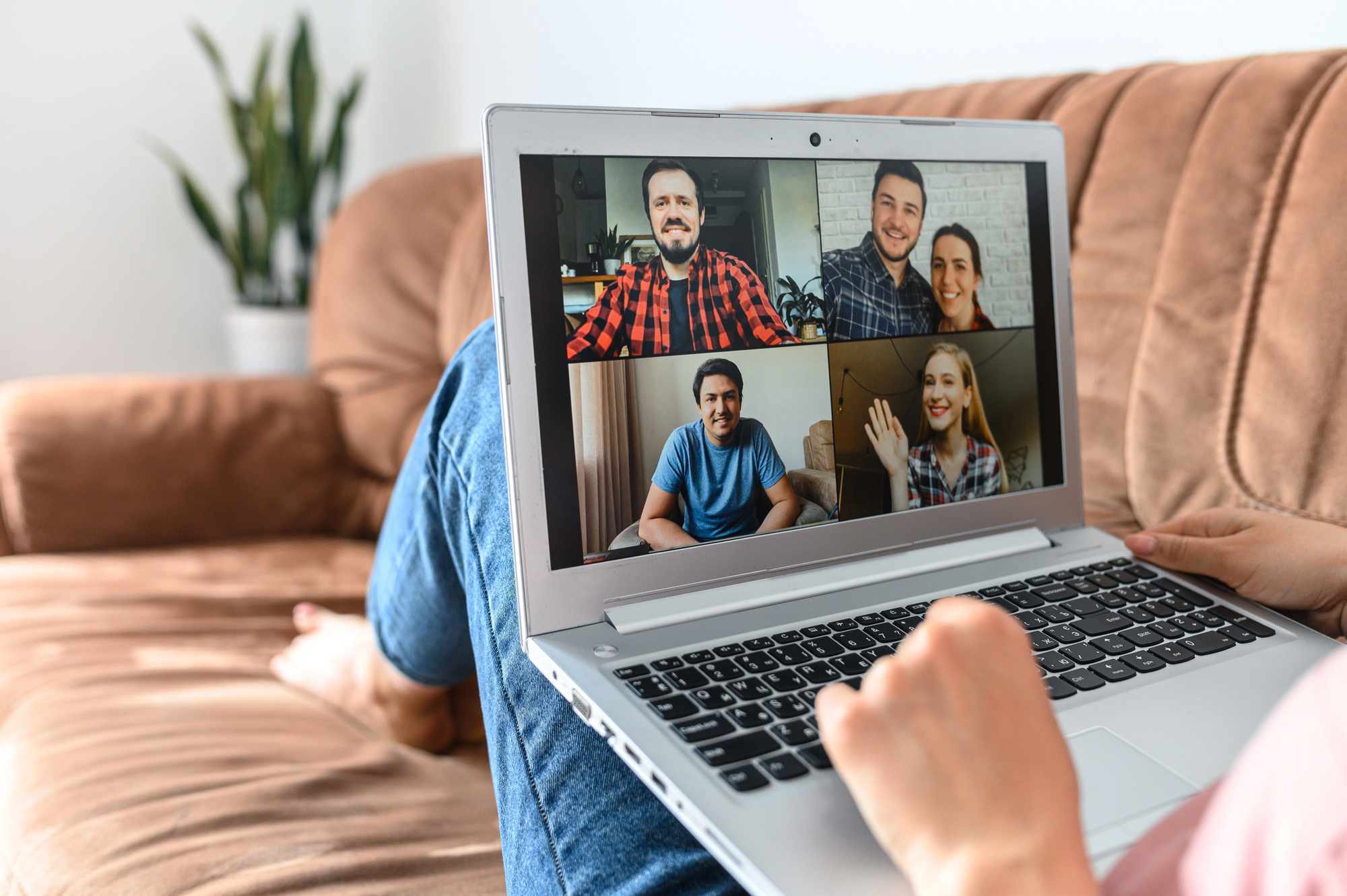 A person sits on a couch, participating in a video call on a laptop. The screen shows four people in separate video boxes. A green plant is visible in the background.