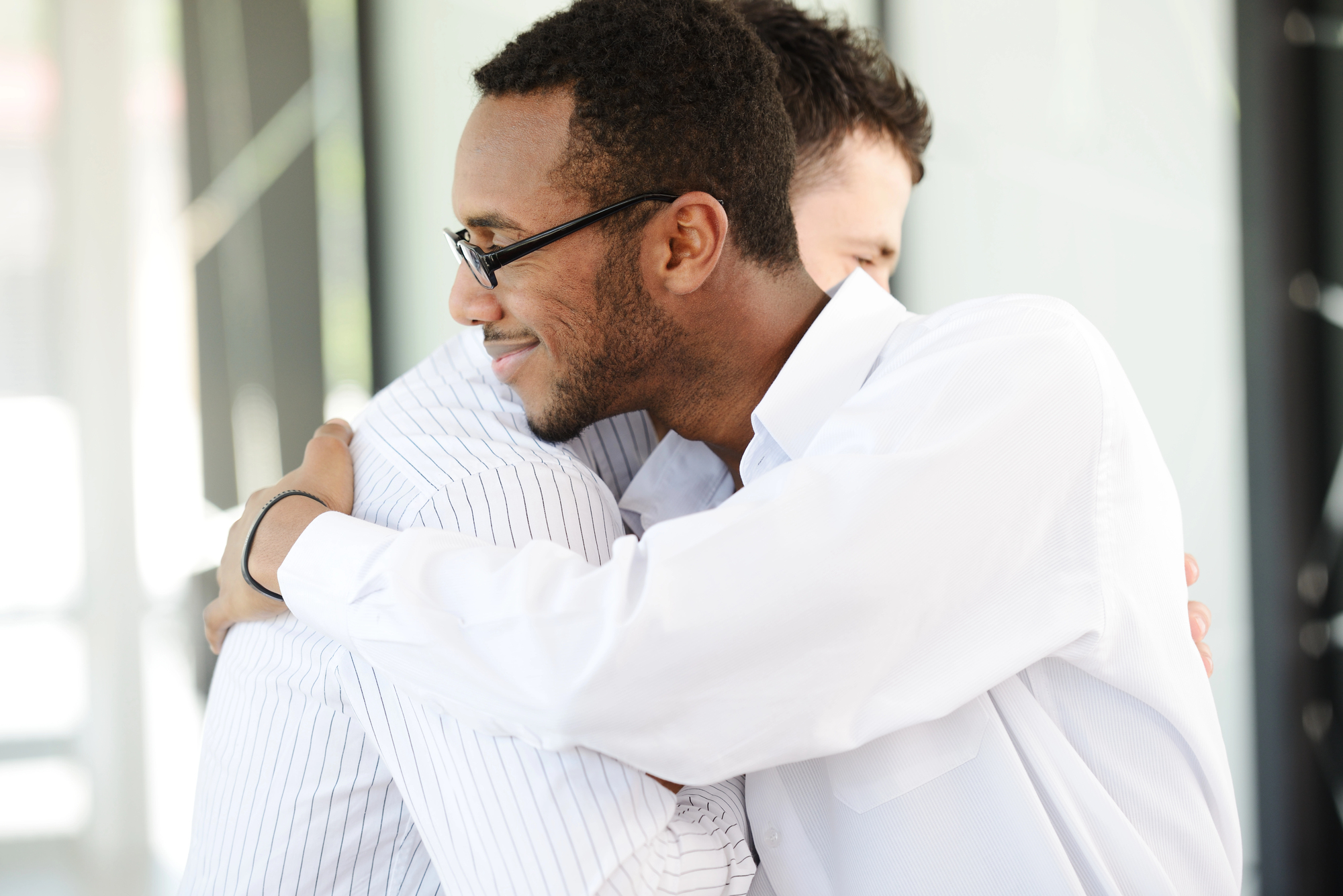 Two men in white shirts embrace warmly. One man wears glasses and a black wristband, smiling as they hug. The background is softly blurred, highlighting their friendly interaction.