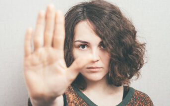 A person with curly hair holds their hand up toward the camera in a gesture that suggests a stop or pause. They are wearing a patterned sweater and standing against a neutral background, looking directly at the viewer.