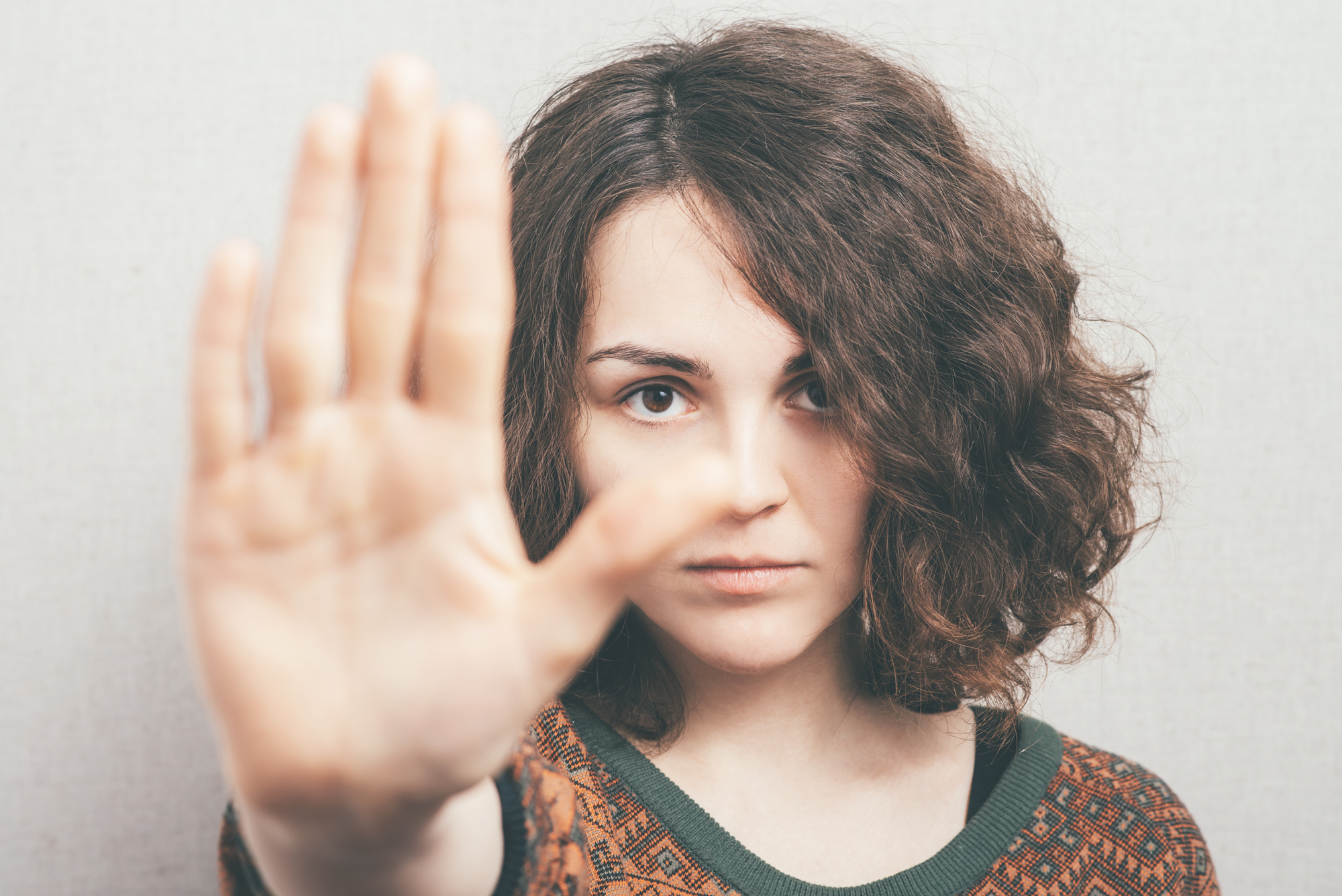 A person with curly hair holds their hand up toward the camera in a gesture that suggests a stop or pause. They are wearing a patterned sweater and standing against a neutral background, looking directly at the viewer.