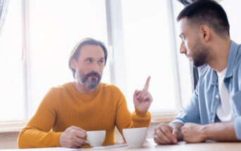 Two men sitting at a table with coffee cups, engaged in conversation. The man on the left, wearing a mustard sweater, raises his index finger, seemingly making a point. Sunlight streams through a window behind them.